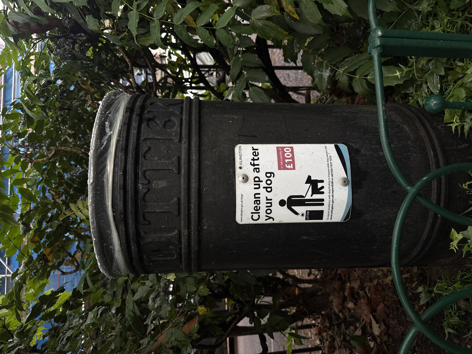 A black trash receptacle stands in a public area, surrounded by lush green foliage. A sign attached to the receptacle reads "Clean up after your dog or risk a penalty of £100" and features an illustration of a person picking up dog waste. The sign also includes the Islington logo, a website address (www.islington.gov.uk/dogs), and a phone number. The receptacle appears to be made of cast iron and has "LITTER" embossed on its side. The area is enclosed by a green metal fence. The scene suggests a park or street in the London borough of Islington, United Kingdom.