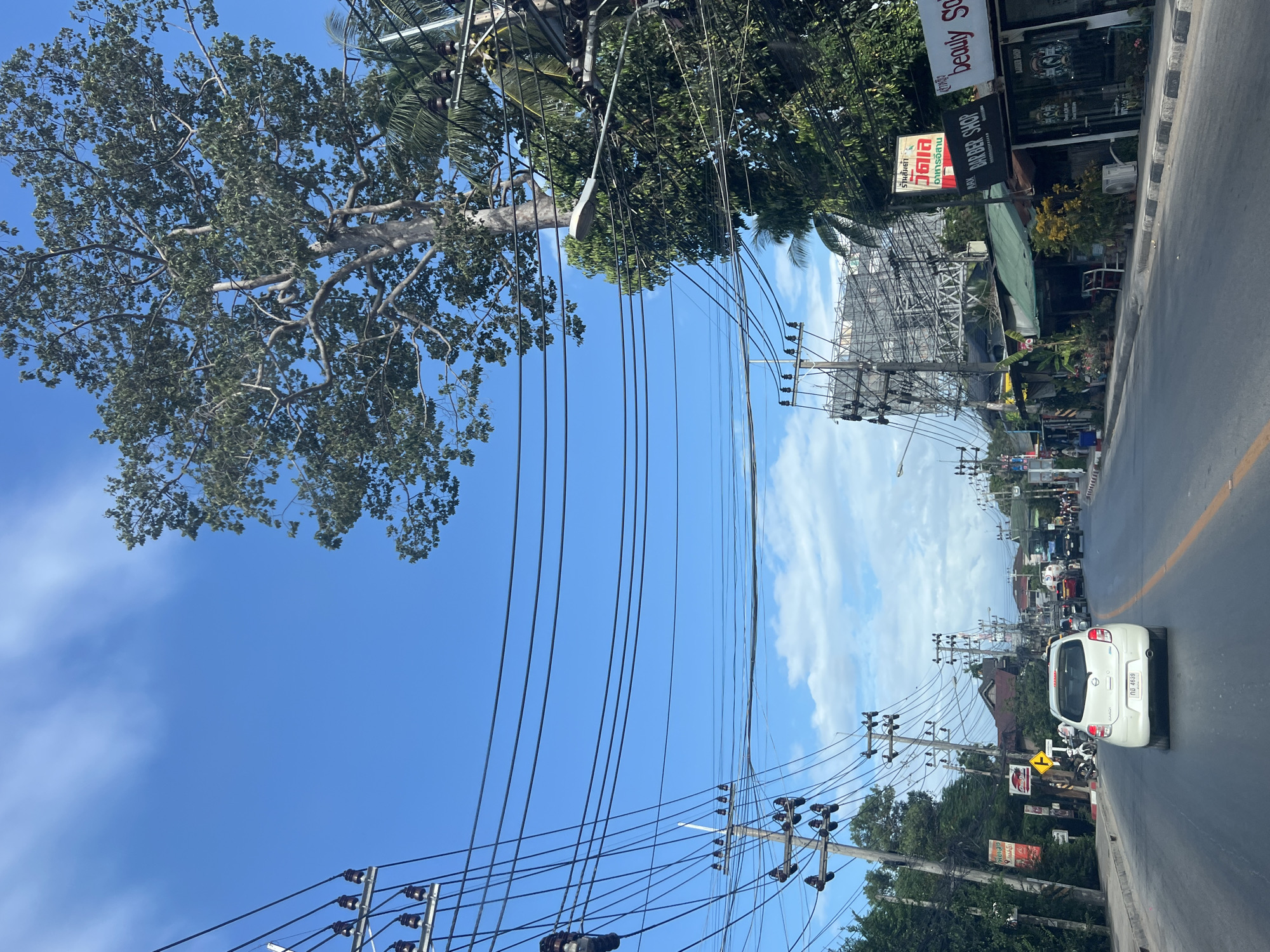 The image shows a street scene in a city. A white car is driving on the road, with other cars and motorcycles in the distance. There are shops and buildings lining the street, as well as utility poles with numerous wires. A large tree is visible in the background. The sky is blue with some clouds.
