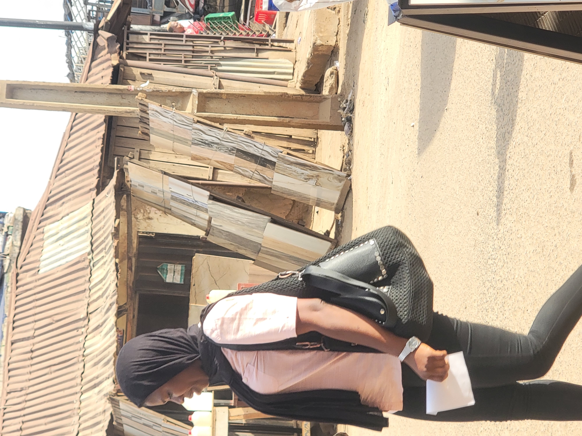 A person wearing a black hijab, a light pink shirt, a black jacket, and black pants is walking on a paved road in Maiburiji, Nigeria. The person is carrying a black shoulder bag and holding a white envelope or paper. The scene is set in a densely populated urban area with buildings constructed from corrugated metal sheets and other materials. Construction materials, including tiles and wooden planks, are visible along the side of the road. The lighting suggests a sunny day.