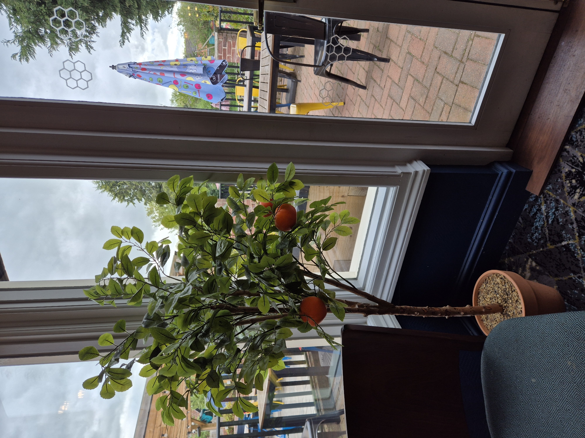 The image captures a scene from indoors, looking out through a large window or glass door into an outdoor seating area. In the foreground, an artificial tree, possibly an orange or citrus variety, stands in a terracotta pot filled with light-colored decorative gravel. The tree has numerous green leaves and a few bright orange, spherical fruits. This potted plant is positioned next to the window, with part of a dark wooden surface and a blue/green patterned armchair visible on the right.
Beyond the window, an outdoor patio or garden with red brick paving is visible under an overcast sky. The outdoor area is furnished with a dark table and several chairs, some black and others a vibrant yellow. A blue parasol with a distinctive, colorful pattern, possibly featuring fruits or drinks, is open and positioned above one of the tables. Further in the background, green trees and some fencing can be seen. Two white, honeycomb-shaped decorations are affixed to the window glass. The scene suggests a pub garden or cafe patio in Needham Market, United Kingdom, during daytime on a cloudy day. No people or visible text are present.