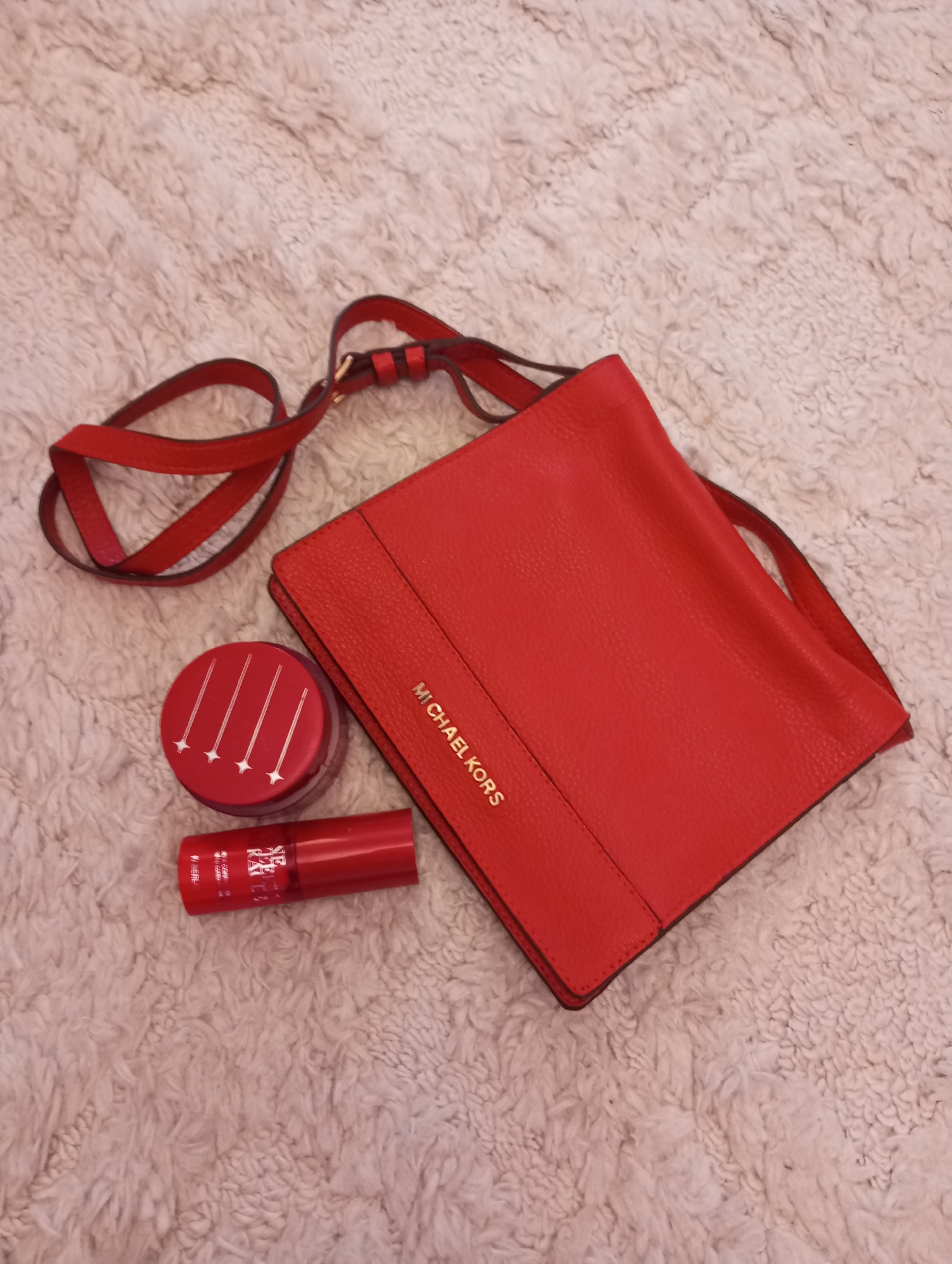 The image shows a red Michael Kors handbag lying on a textured, light-colored rug. Beside the handbag, there is a round container with white line patterns and a short, red tube. The location in the city of Chaikovsky, Russia is not determinable from the image.