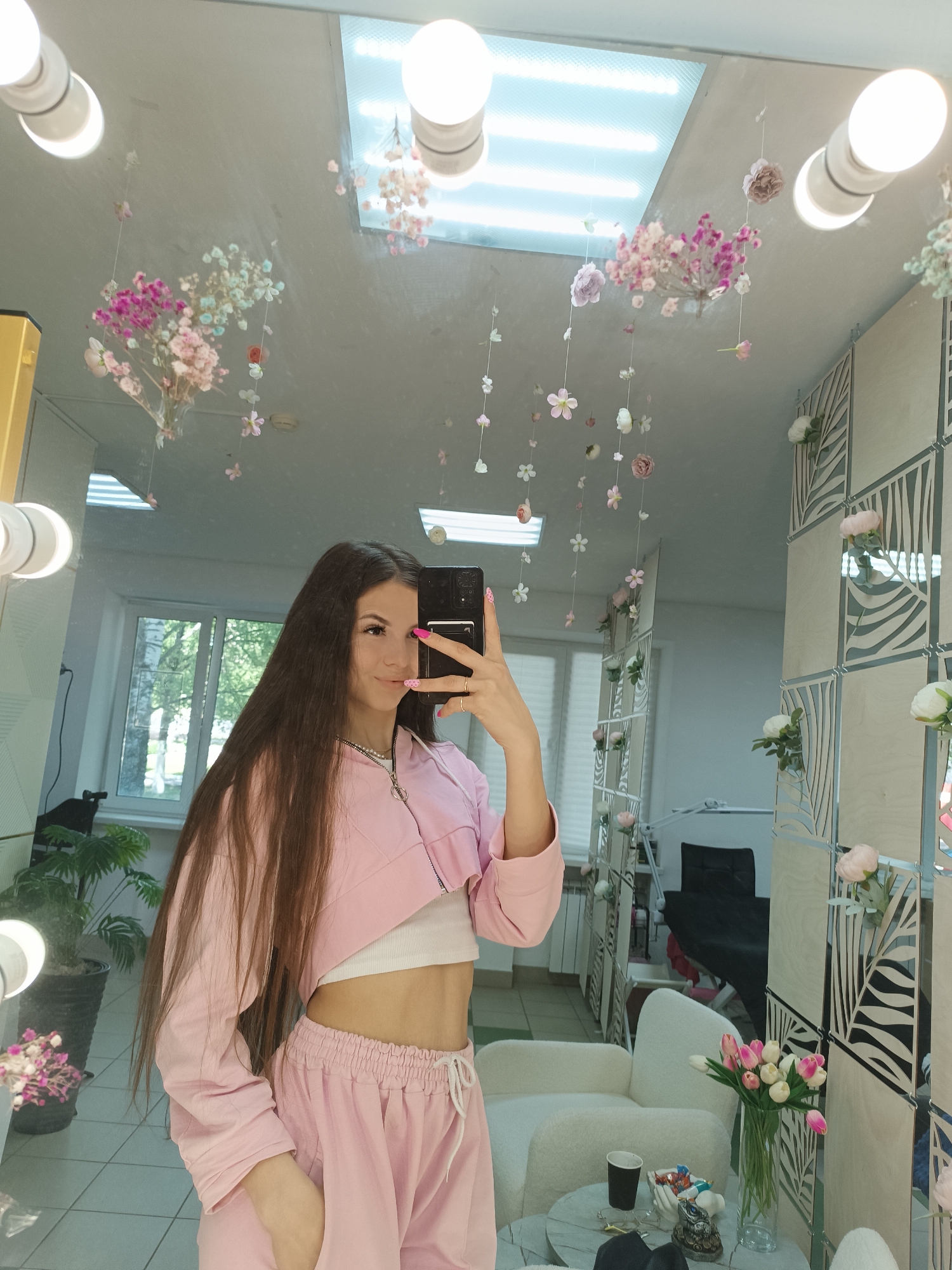 A full-length reflection of a woman holding a smartphone is visible in a mirror. She is wearing a pink tracksuit, consisting of a cropped jacket and drawstring pants, and a white top, revealing her midriff. Her hair is long and brown. The mirror is framed with light bulbs. Hanging from the ceiling above are artificial floral arrangements. The reflection also shows a room with a window, a plant, a white armchair, a small table with items on it, a vase of tulips, and a folding screen decorated with foliage. The location is unknown.