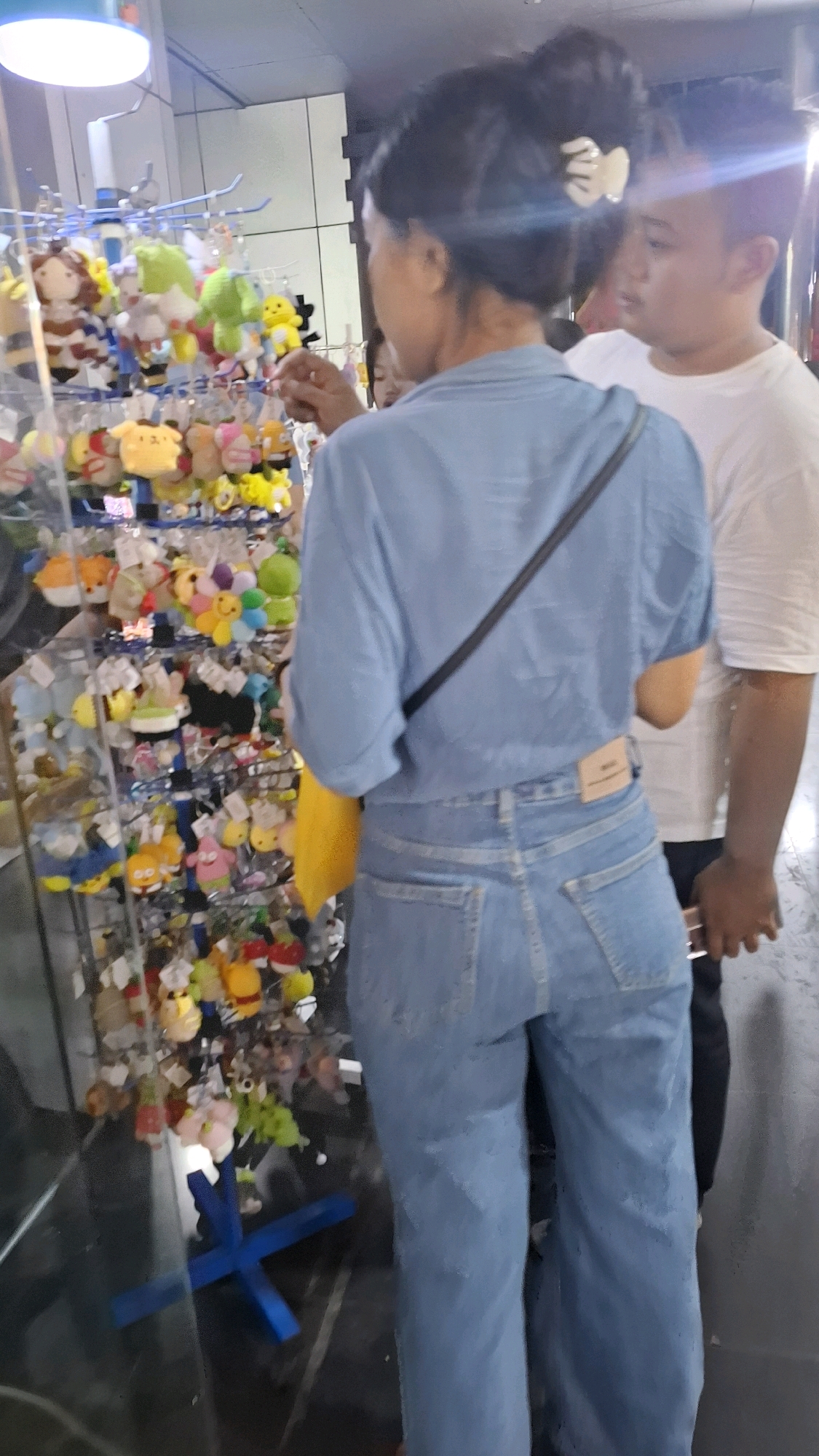 A woman with her hair in a bun and a white clip is examining a display of plush toys in a store. She is wearing a light blue button-down shirt and light blue denim jeans. A yellow bag is tucked under her arm, and a dark strap goes across her chest. To her right, a man wearing a white t-shirt is looking forward. The plush toys are displayed on multiple tiers of a rack. In the background, a white tiled wall is visible. The scene appears to be in an indoor retail environment. There is no information available to determine the specific location within the city of Bojonegoro, Indonesia.