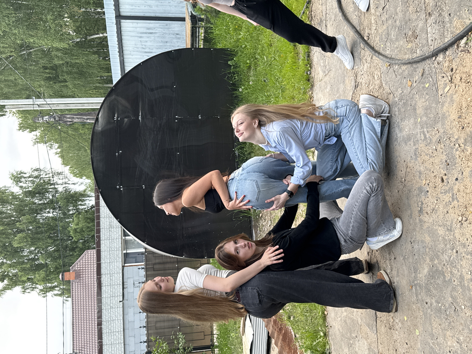 The image shows four young women in an outdoor setting, likely during the daytime on a cloudy day. They are positioned in a pyramid-like structure, with three of them forming a base and one on top. The woman at the bottom of the pyramid, wearing a white t-shirt and dark jeans, is lying on her side. On top of her are two other women: one in a black top and dark jeans is sitting with her legs extended, and another in a light blue shirt and light blue jeans is kneeling and leaning on the woman in black. The fourth woman, wearing a black top and dark jeans, is positioned at the top of the pyramid, kneeling with her legs bent. Her hands are on the shoulders of the woman below her.
The background features a large, dark, circular object, possibly a satellite dish, against a backdrop of trees and buildings. The ground is a mix of concrete and dirt. The overall mood appears casual and playful, with the women engaged in a cooperative activity. The location context provided indicates Perm, Russia. There is no visible text in the image.