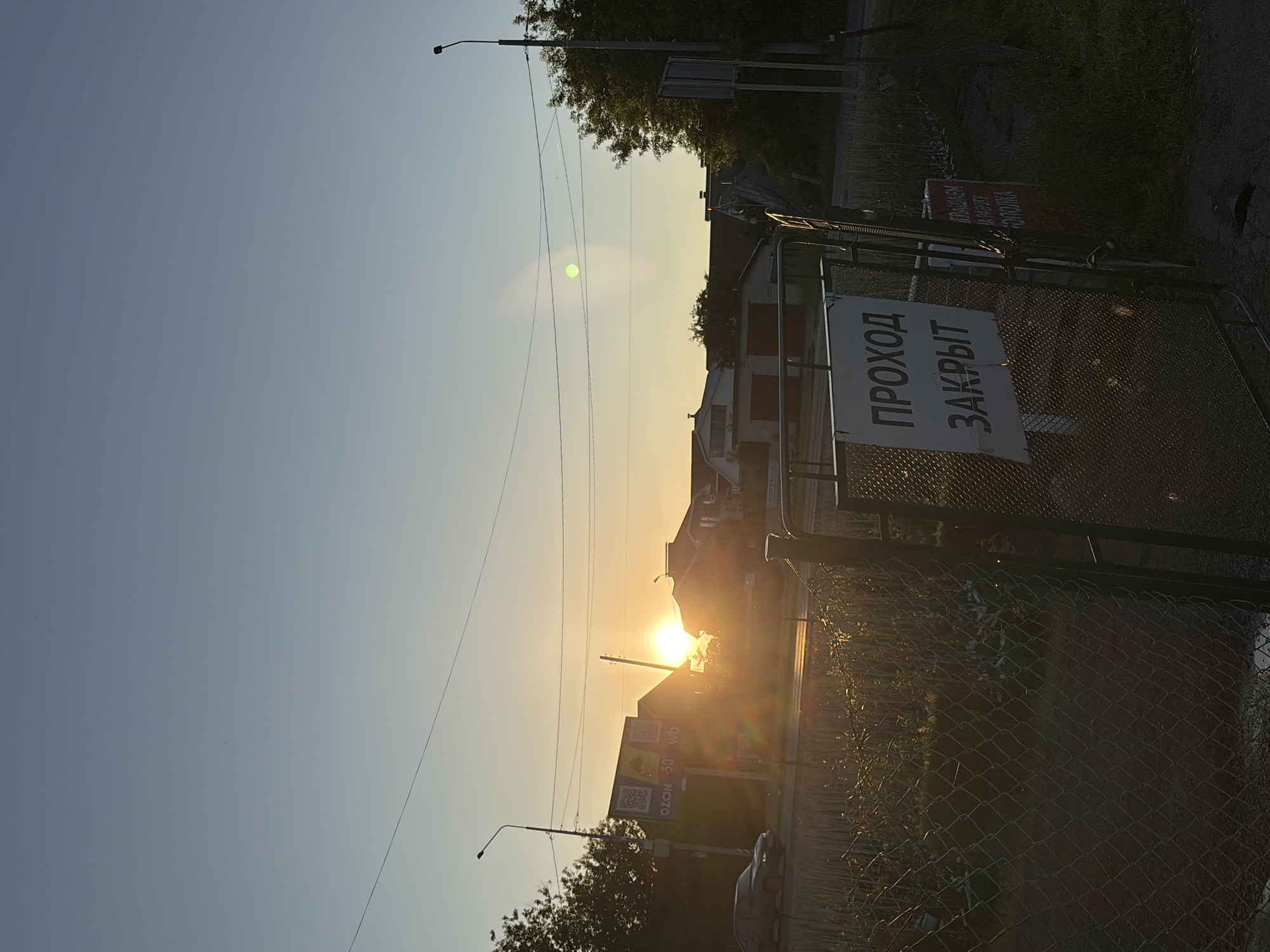 The image captures an outdoor scene at sunset, in an unknown location characterized by Russian text on signs. The sky, a gradient from light blue at the top to warm yellow-orange at the horizon, dominates the upper frame. The sun is visible as a bright, circular disc on the horizon, centered horizontally, and slightly left of the vertical center, casting a strong lens flare, including a prominent green circular artifact in the mid-upper right sky.
In the foreground, a dark metal fence, possibly chain-link, runs across the bottom of the frame. Attached to a gate in the fence is a rectangular white sign with dark text, which reads "ПРОХОД ЗАКРЫТ" (Passage Closed) in Cyrillic. To the right of this sign, a smaller, reddish sign is partially visible, displaying text that includes "ПРИГЛАШАЕМ НА РАБОТУ" (Inviting to work). The area immediately behind the fence appears to be overgrown with dark foliage and some debris.
The midground features a paved road or street, reflecting the sun's glare. A dark-colored sedan is visible on the left side of the road, moving away from the viewer. Several utility poles with overhead electrical wires crisscross the frame horizontally. On the left, a billboard displays QR codes and text, including "OZON -50 wb," indicating commercial advertisements. Trees with green foliage are present on both sides of the road, partially silhouetted.
In the background, multiple residential houses with varying rooflines and architectural styles are visible, mostly silhouetted against the bright sunset. No people are discernible in the scene. The overall lighting suggests late afternoon or early evening.