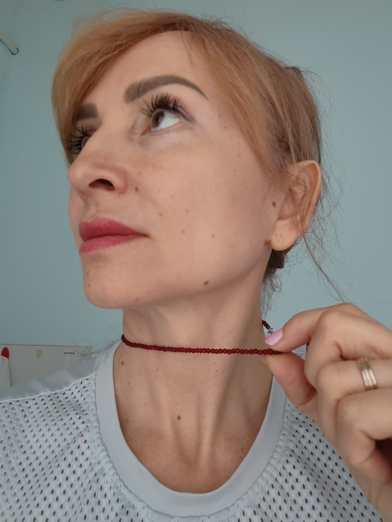 The image depicts a female subject, positioned from the mid-chest upward, angled slightly to her left, looking upwards. She has light reddish-brown hair, styled with bangs, and fair skin with visible freckles and moles. Her eyes feature extended false eyelashes, and her lips are adorned with dark pink lipstick. She wears a light-colored mesh-patterned top and a thin, dark red beaded choker around her neck. Her left hand, featuring a light pink manicured nail on the index finger and a gold-toned multi-band ring on the ring finger, is shown gently pinching the red choker near her clavicle. The background consists of a plain, light blue wall. In the lower-left quadrant, a light-colored object with faint Cyrillic text, possibly "жизнь как," is visible, along with a small red shape. The scene is located in Chaikovsky, Russia.