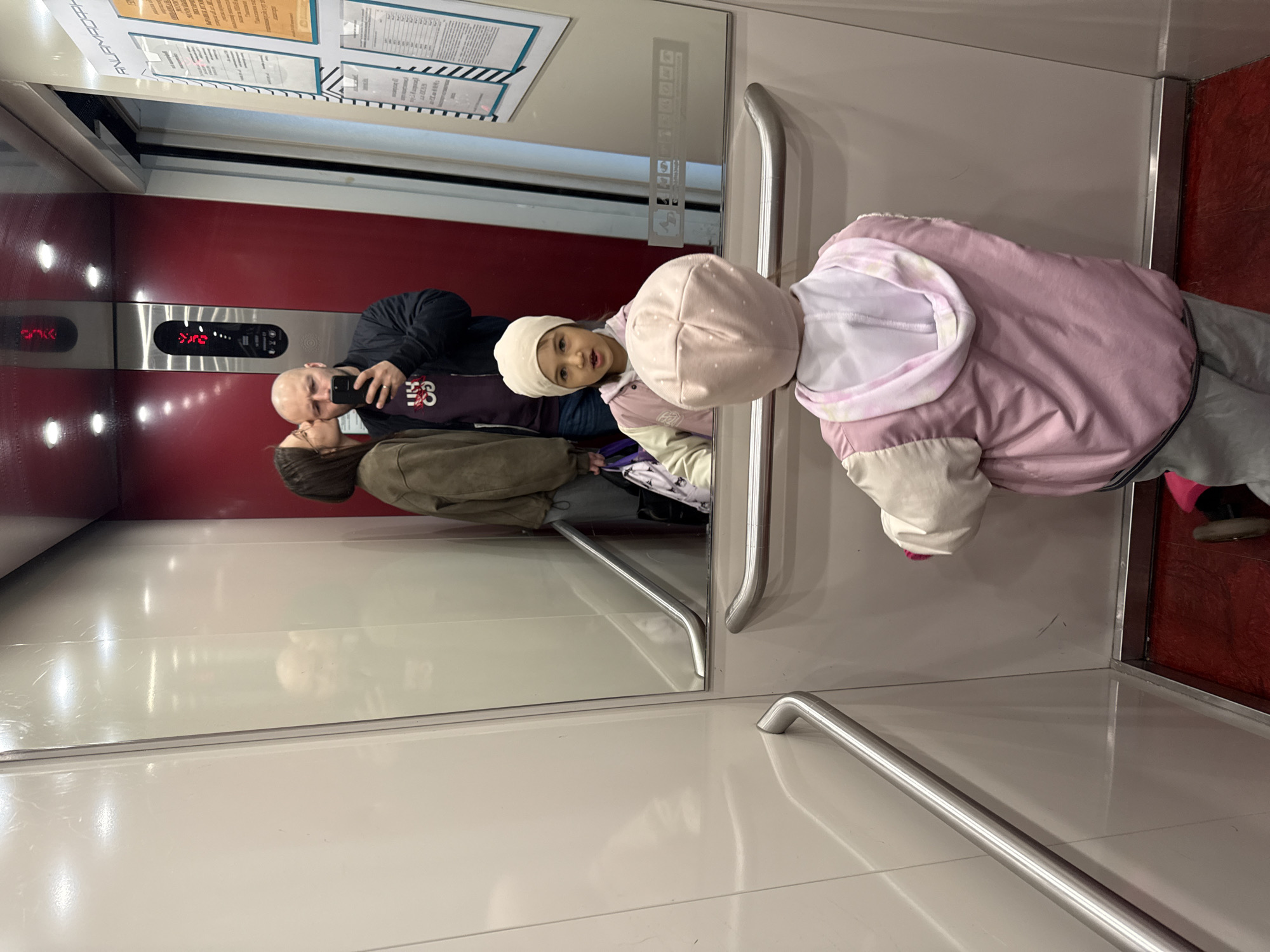 The image captures a family inside an elevator. The elevator has red interior paneling on the back wall and white side panels. A display above shows the current floor number as 276, and control buttons are located below. A man, presumably the father, is taking a selfie with his mobile phone. He is bald and wearing a dark jacket over a maroon shirt. A woman with brown hair, likely the mother, stands beside him wearing an olive green jacket. A young girl wearing a white beanie and a pink hooded jacket stands in front of them. The elevator appears to have a mirror, and the photo is taken from that angle. A silver handrail is visible on the side of the elevator. The image was taken in Krasnoyarsk, Russia, as indicated by the Russian language sign visible in the mirror.