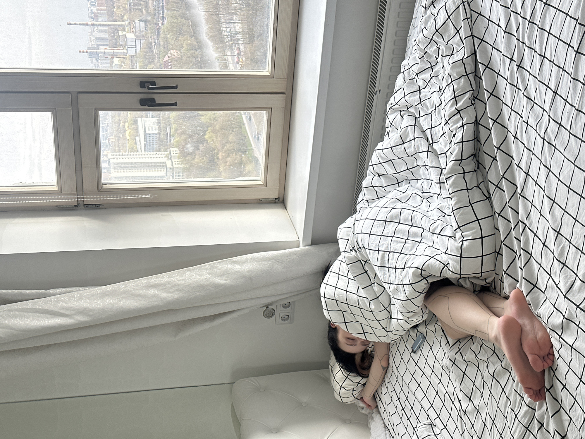 This media file captures an indoor scene, likely a bedroom in Khimki, Russia, presented with a 90-degree clockwise rotation. A person is seen sleeping or resting in a bed, largely covered by a white duvet with a prominent black grid pattern. The person's face is partially visible, nestled into the covers, while their feet and legs, adorned with a distinctive geometric line pattern (possibly a tattoo or body art), protrude from the bottom right of the duvet.
A large window with a light-colored wooden frame dominates the left side of the image, allowing ample natural light into the room. Through the window, an urban landscape is visible, featuring various buildings, some with industrial chimneys, sparse trees, and a road. The sky appears overcast or hazy. White window blinds are partially visible at the base of the window frame, and a white window sill runs beneath it.
To the right of the bed, a white, ribbed radiator is positioned against a wall. Near the head of the bed, on the wall, two European-style electrical outlets are visible alongside a small dial or switch. A light-colored curtain is drawn to the side. The bed has a white, possibly upholstered headboard with tufting. The scene suggests a peaceful daytime moment of rest or sleep.