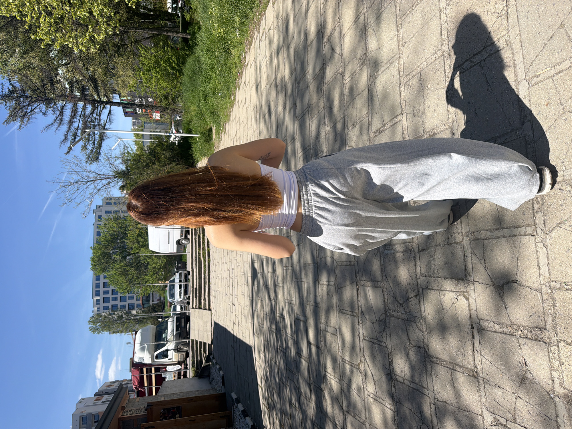 A woman with long red hair, wearing a white top and grey sweatpants, stands on a light grey brick paved sidewalk. The sidewalk is shaded by trees, with dappled sunlight creating patterns. To the left of the sidewalk is a grassy area, trees, buildings, and parked white vans. The sky is blue. The scene is in Sofia, Bulgaria.