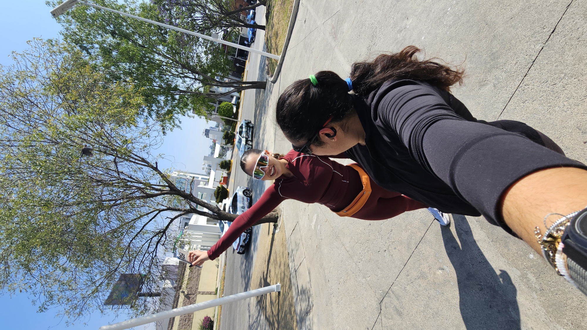Two women pose for a selfie on a sidewalk in Puebla, Mexico. One woman, on the left, wears sunglasses, a burgundy athletic outfit, and a yellow fanny pack. She extends her arm towards the camera and smiles. The second woman, partially visible on the right, wears a black long-sleeved shirt. She has her hair pulled back with a green hair tie. A street with parked cars and buildings is behind them. A tree with green leaves stands to the left of the women. The sky is bright blue.
