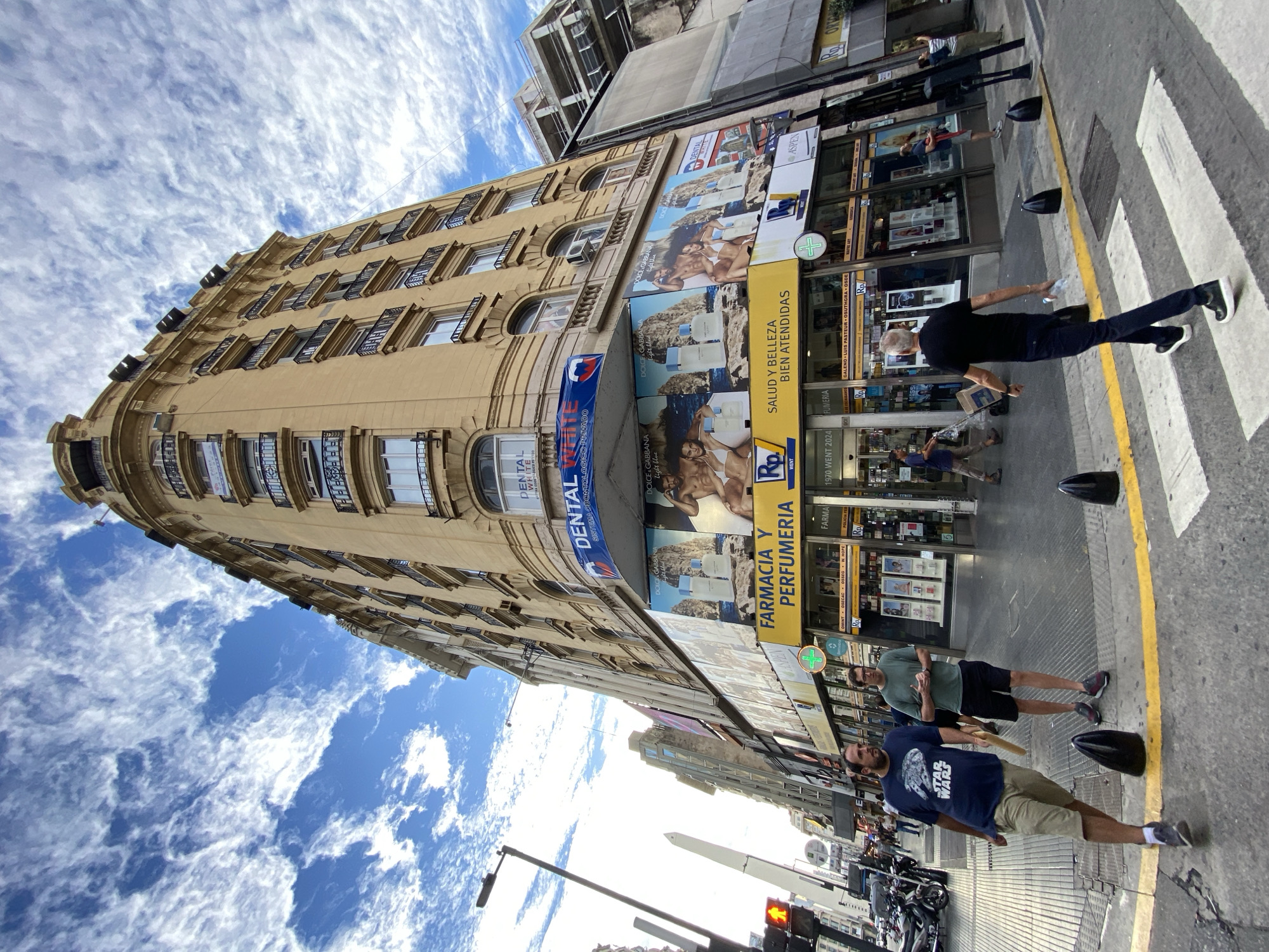 The image captures a street scene in Buenos Aires, Argentina, with a focus on a multistory building. The building, characterized by its light-colored facade and architectural details, features multiple windows with dark shutters. Advertisements, including those for fragrances and dental services, are prominently displayed on the building's exterior, alongside signage for a "Farmacia y Perfumeria" and other businesses. Pedestrians are present on the sidewalk, including individuals crossing a zebra crossing. Motorbikes are parked along the street. The sky above is blue with scattered clouds. A traffic light is visible, adding to the urban environment.