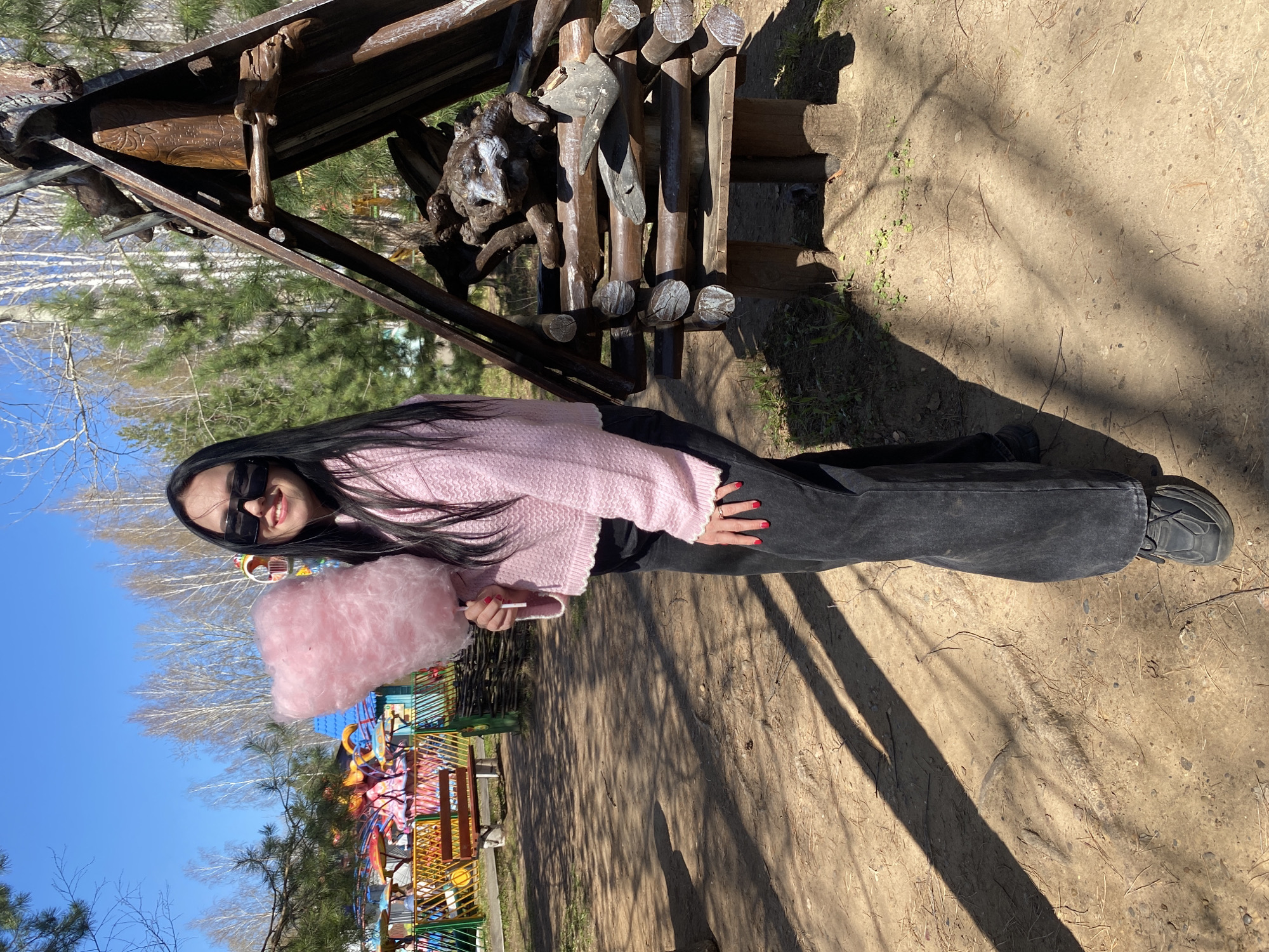 A young woman stands in a park in Chaikovsky, Russia, holding pink cotton candy. She wears a pink sweater, black pants, and black sneakers. She is also wearing sunglasses. Behind her is a colorful playground and some trees. To her right, a wooden structure sits on dirt ground. The sky is blue.