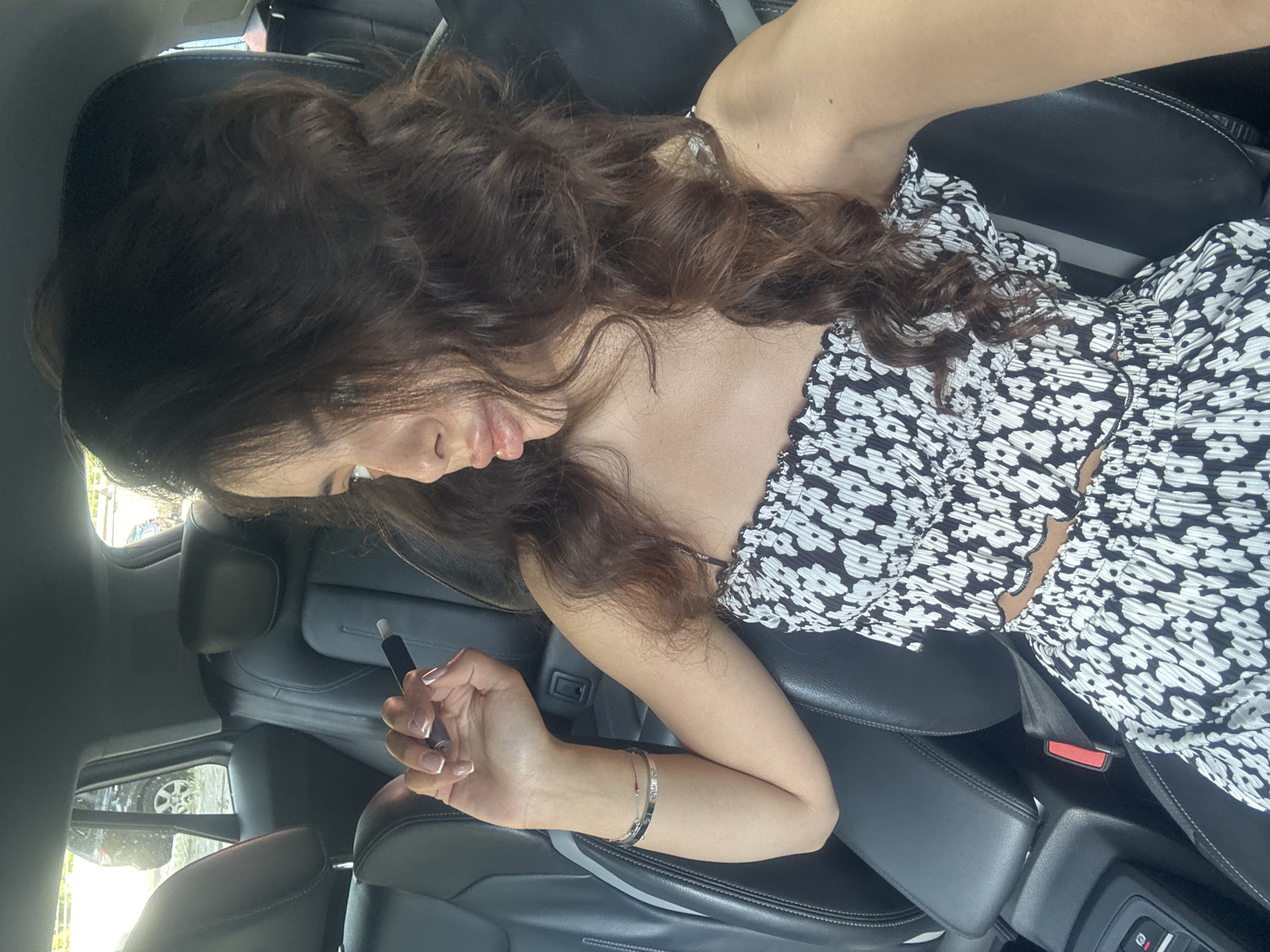 A young woman with long, dark, wavy hair is seated in the back seat of a car. She is wearing a black and white floral print two-piece outfit. Her right arm is raised, and she is holding a black object, possibly an electronic cigarette, between her fingers. She is looking away from the camera to the right. The car interior is visible, with black leather seats and the central console. The background outside the car windows appears to be a street scene, possibly in Plovdiv, Bulgaria, with other vehicles and trees visible.