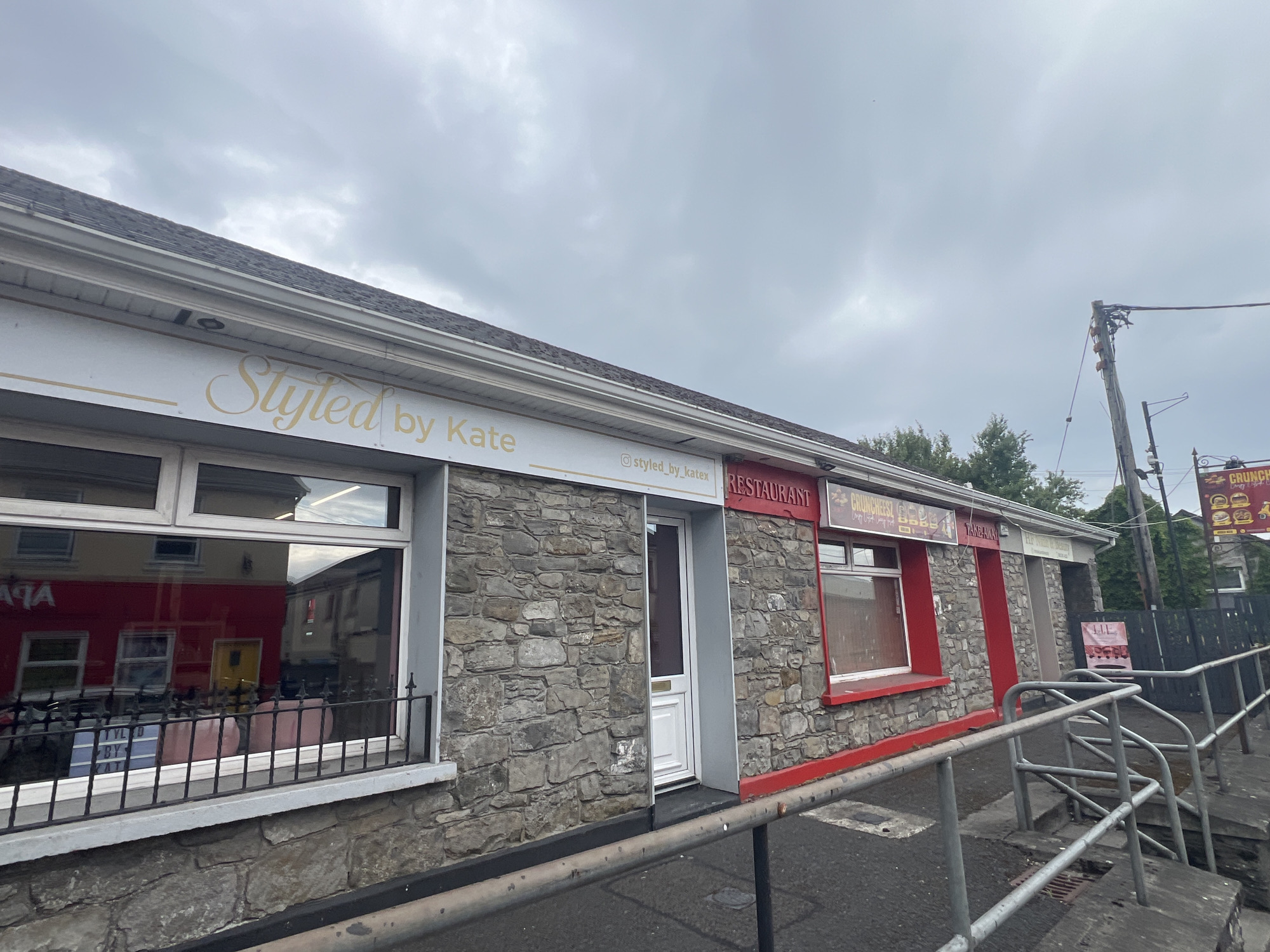 The image shows the exterior of a small commercial building. The facade is constructed of rough-hewn grey stone. A white awning runs along the top of the building, with gold lettering that reads "Styled by Kate". Below this, there is a window on the left and a door on the right. The window is reflective and shows the interior of the establishment, which appears to be a salon with pink chairs and a lighted sign with text. To the right of the door, there is a red sign that says "RESTAURANT" and another sign for "CRUNCHES T". The building is situated on a street with a grey metal handrail and steps leading up to the entrance. The sky is overcast with grey clouds.