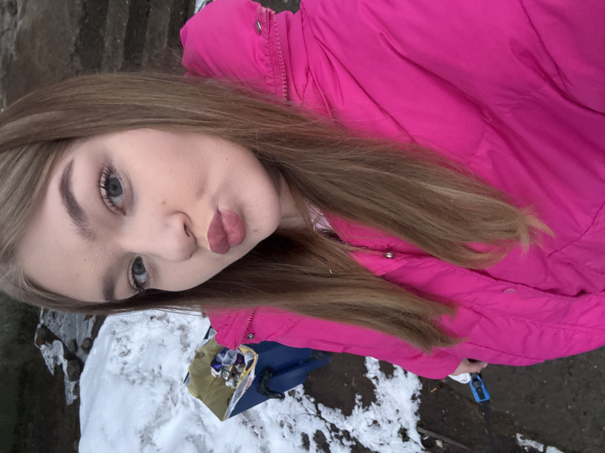 A young woman with light brown hair and blue eyes, wearing a bright pink winter jacket, poses for a selfie. Her lips are puckered in a kissing expression. The background shows a snowy area mixed with bare ground, possibly a park or a roadside location. There is a glimpse of a blue object and some discarded items in the snow. Based on the prompt, this scene may be in the city of Chaikovsky, Russia.