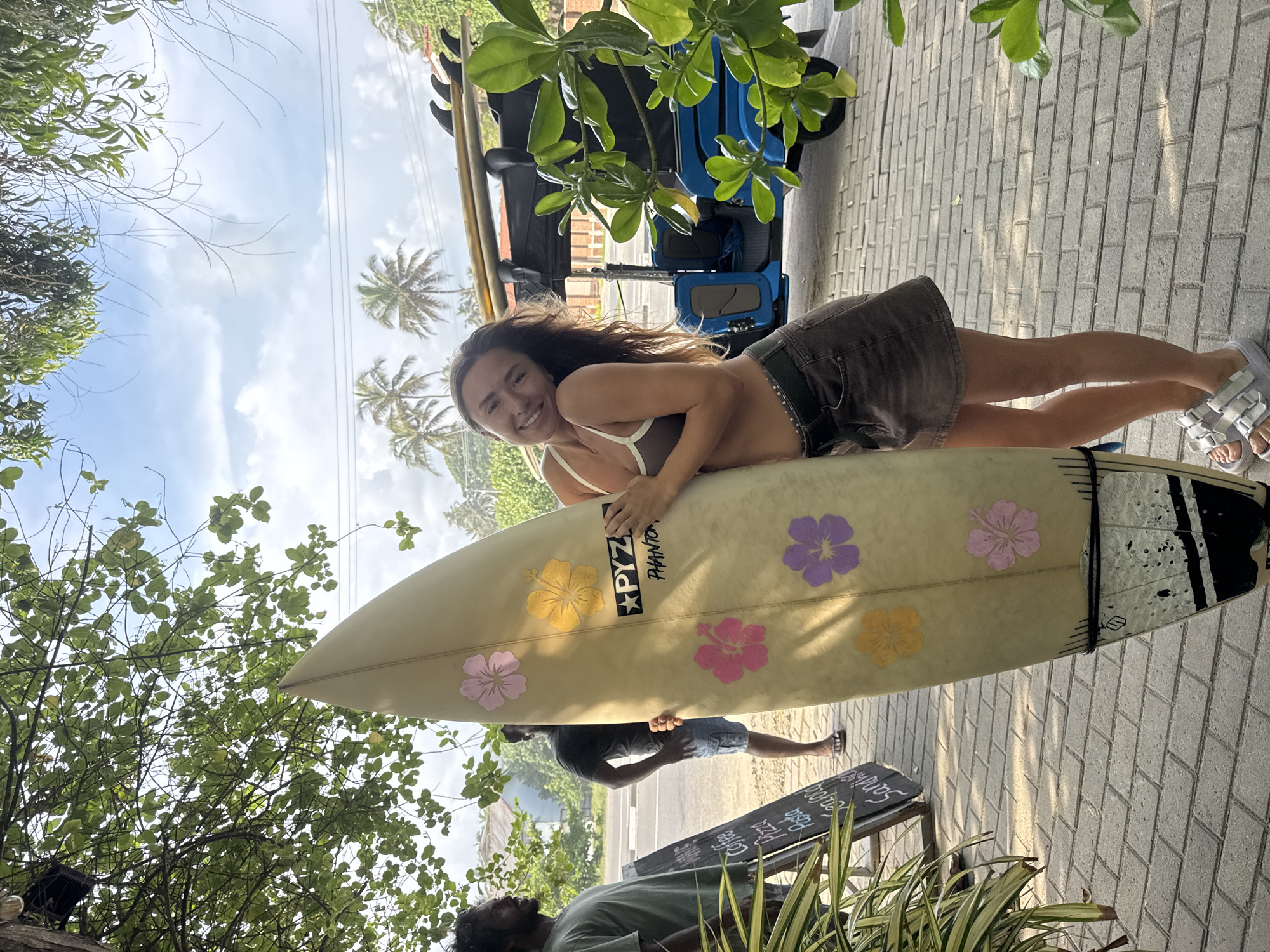 The image depicts a young woman lying on a surfboard adorned with hibiscus flower designs. The surfboard leans against a building and extends into a sidewalk area in Kathaluwa, Sri Lanka. The woman is smiling, wearing a brown bikini top and a dark skirt. Her sandals are white. A blue tuk-tuk is parked in the background along with other surfboards. The scene is filled with tropical foliage, including palm trees and other greenery. Two men are standing near a menu stand that says "Pizza & Coffee." The area is paved with gray brick. The sky is partly cloudy.