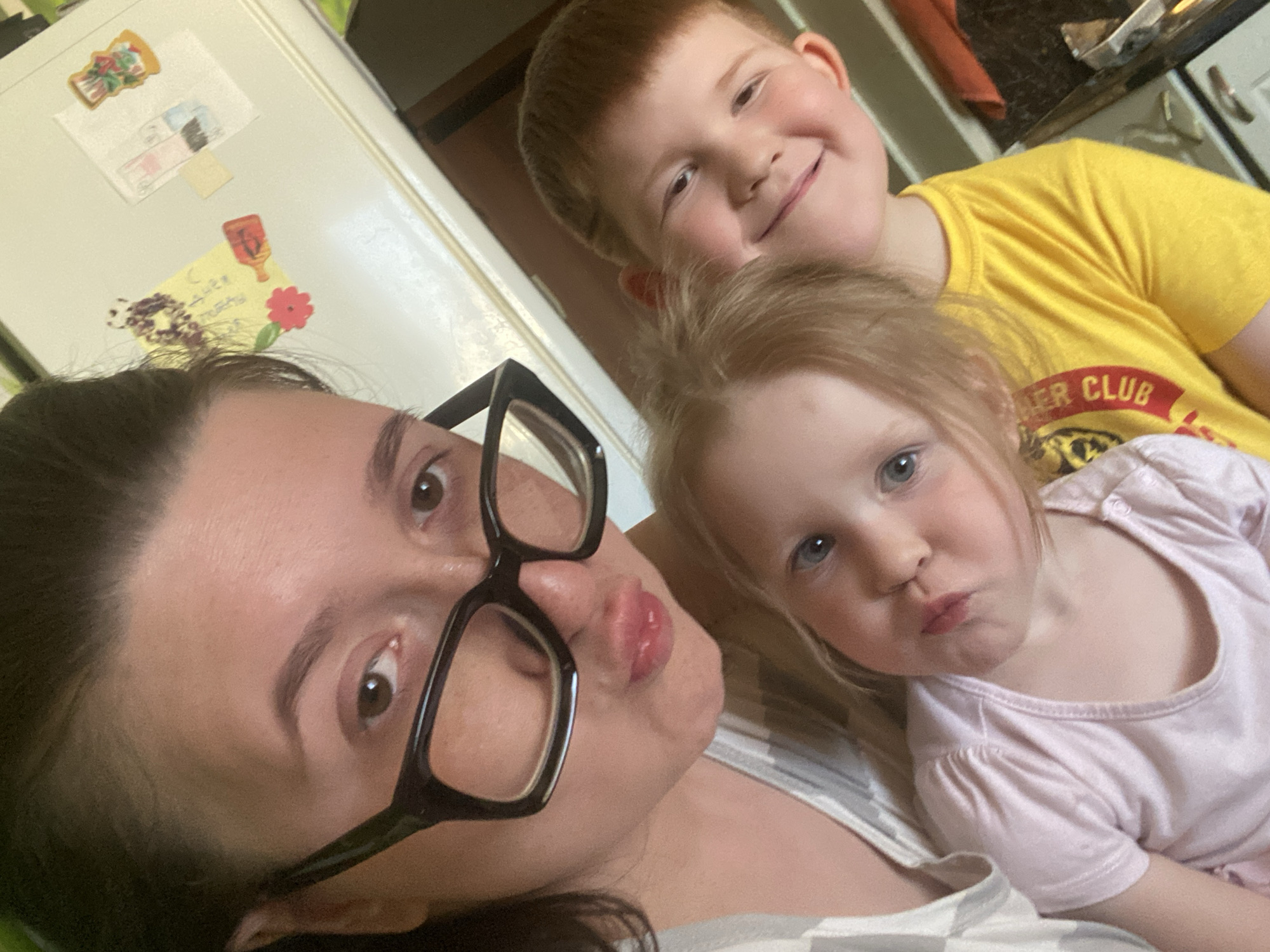 A selfie shows a woman wearing large, black-framed glasses, positioned in the foreground and left of the frame. She is making a "duck face." Behind her and to the right, there are two children, a boy and a girl. The boy, wearing a yellow t-shirt with a "Miller Club" logo, is smiling. The girl, wearing a light pink shirt, is also making a "duck face." They appear to be indoors. The background shows a white refrigerator covered with magnets and drawings, and part of a kitchen counter. The location is unknown and there is no evidence in the photo that suggests it is in Chaikovsky, Russia.