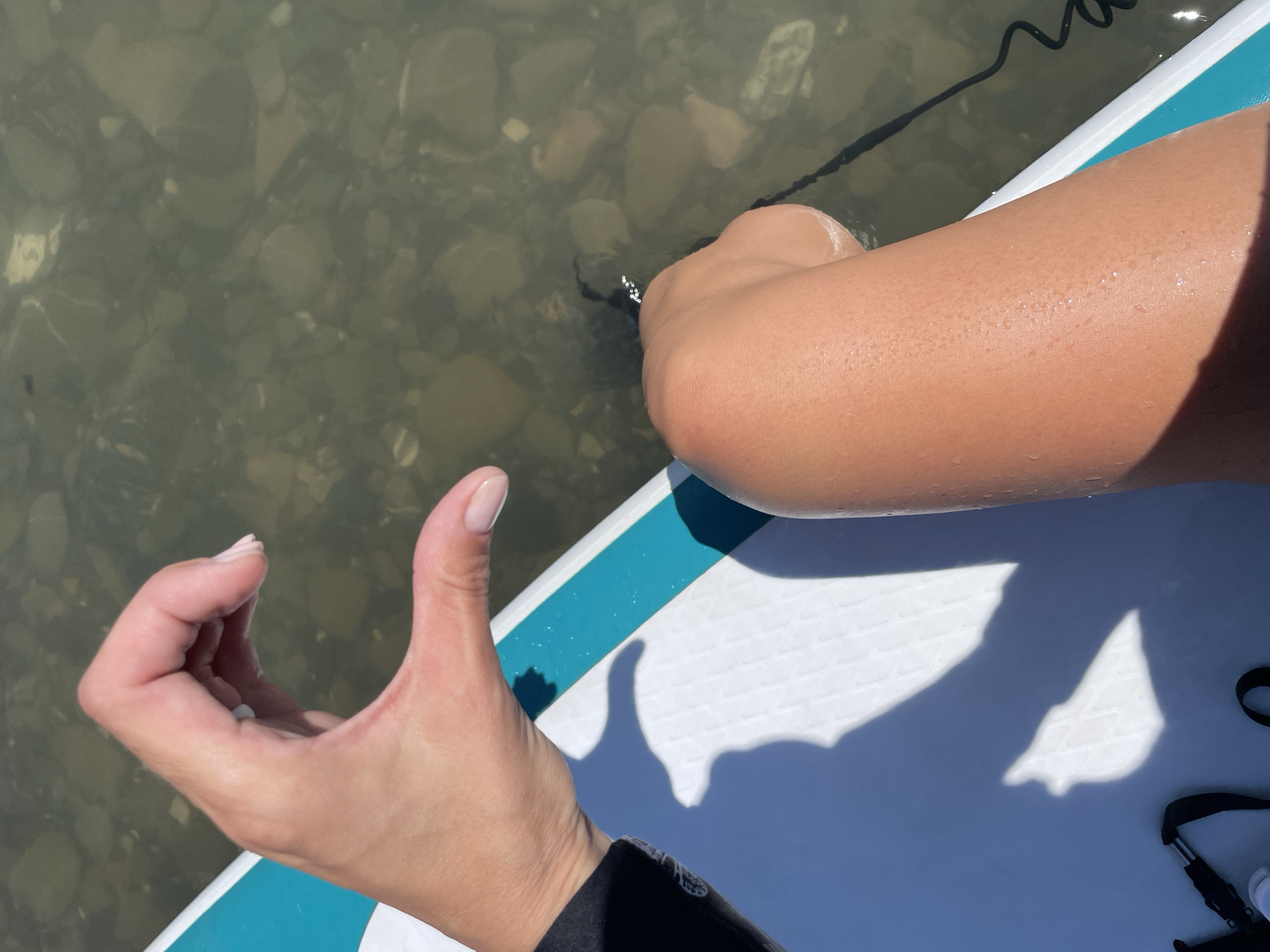 The image captures a first-person perspective of a person engaging in a water activity, likely paddleboarding, in Sochi, Russia. The scene features a person's sun-kissed arm, hand, and leg, along with a white and teal paddleboard. The individual's right leg is partially submerged in shallow, clear water, revealing smooth, light-colored pebbles and rocks on the seabed beneath. A dark leash or rope, possibly connected to the paddleboard, is visible in the water near the leg.
Water droplets glisten on the person's skin, indicating recent contact with the water. The left hand, wearing a dark sleeve, is giving a "thumbs up" gesture, suggesting a positive experience or approval. Strong sunlight is evident, casting distinct shadows of the person's body and hand onto the surface of the paddleboard. The paddleboard itself has a textured, non-slip surface. The lighting indicates a bright, sunny day. The clear water over the rocky bottom suggests a calm sea or lake environment. There is no visible text in the image.