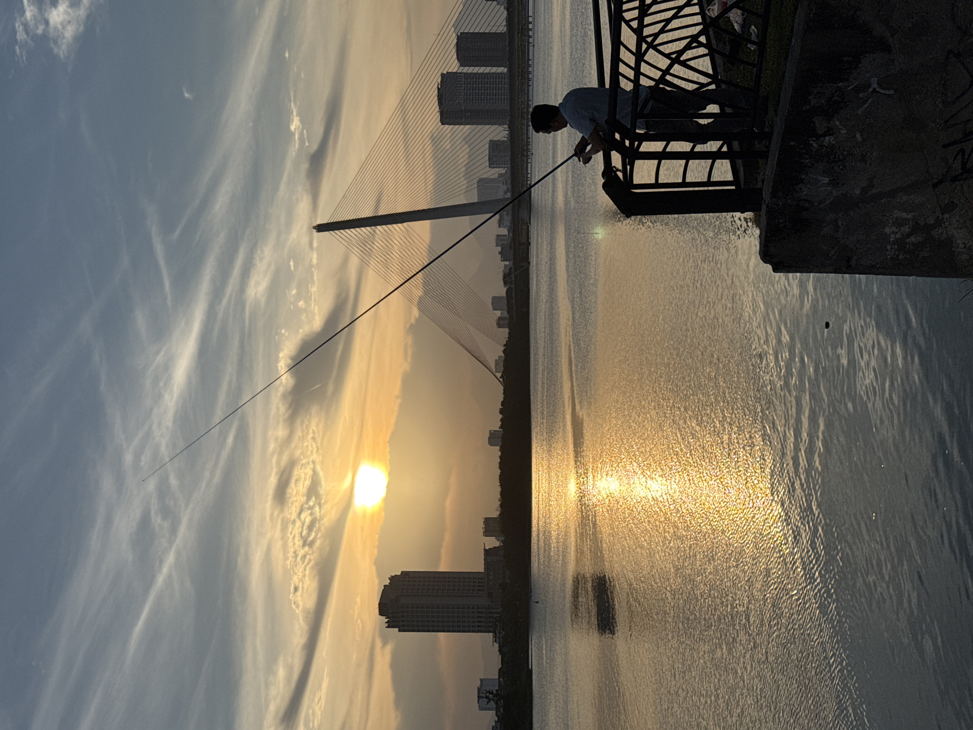 A person fishes off a promenade railing at sunset in Da Nang, Vietnam. The sun is low on the horizon, casting a golden glow on the water and sky. Tall buildings and a cable-stayed bridge are visible in the distance. The scene is serene and peaceful, with the gentle ripple of water and the silhouette of the fisherman creating a sense of calm.