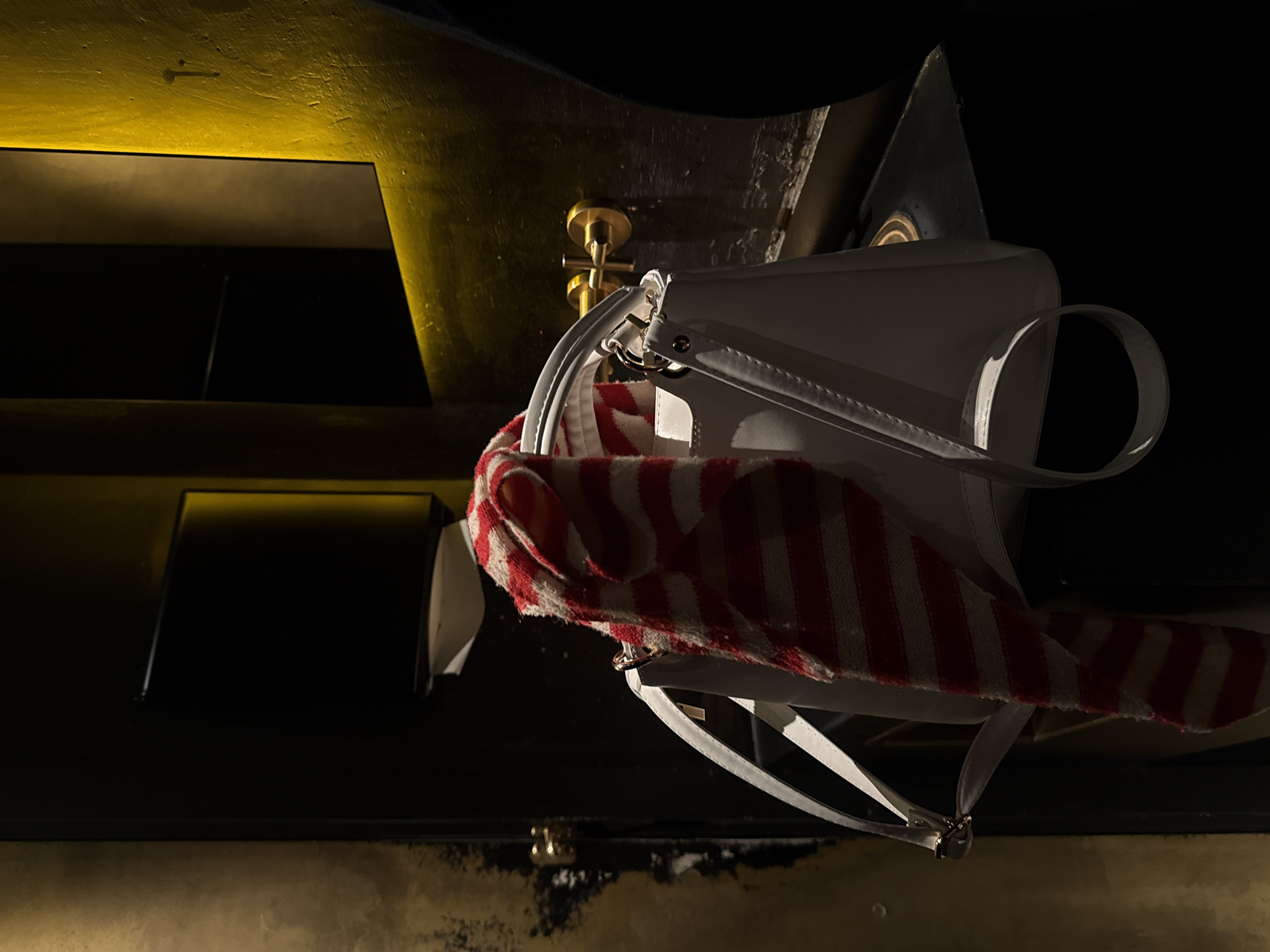 The image depicts a dimly lit indoor scene, likely a public restroom. Centrally positioned is a white, rigid handbag with a red and off-white striped textile draped over its top handles and extending down its front. In the background, a dark, non-reflective wall features a golden-toned wall-mounted faucet with a cross-shaped handle. To the left of the faucet, a black paper towel dispenser contains a single white paper towel. A rectangular mirror, appearing dark, is partially visible above and to the right of the faucet. An unseen light source emits a warm yellow glow along the top edge of the mirror and onto an adjacent textured wall, which displays mottled brown and gold tones. The specific location within Yerevan, Armenia, is not identifiable from the image. No individuals are present in the scene.
