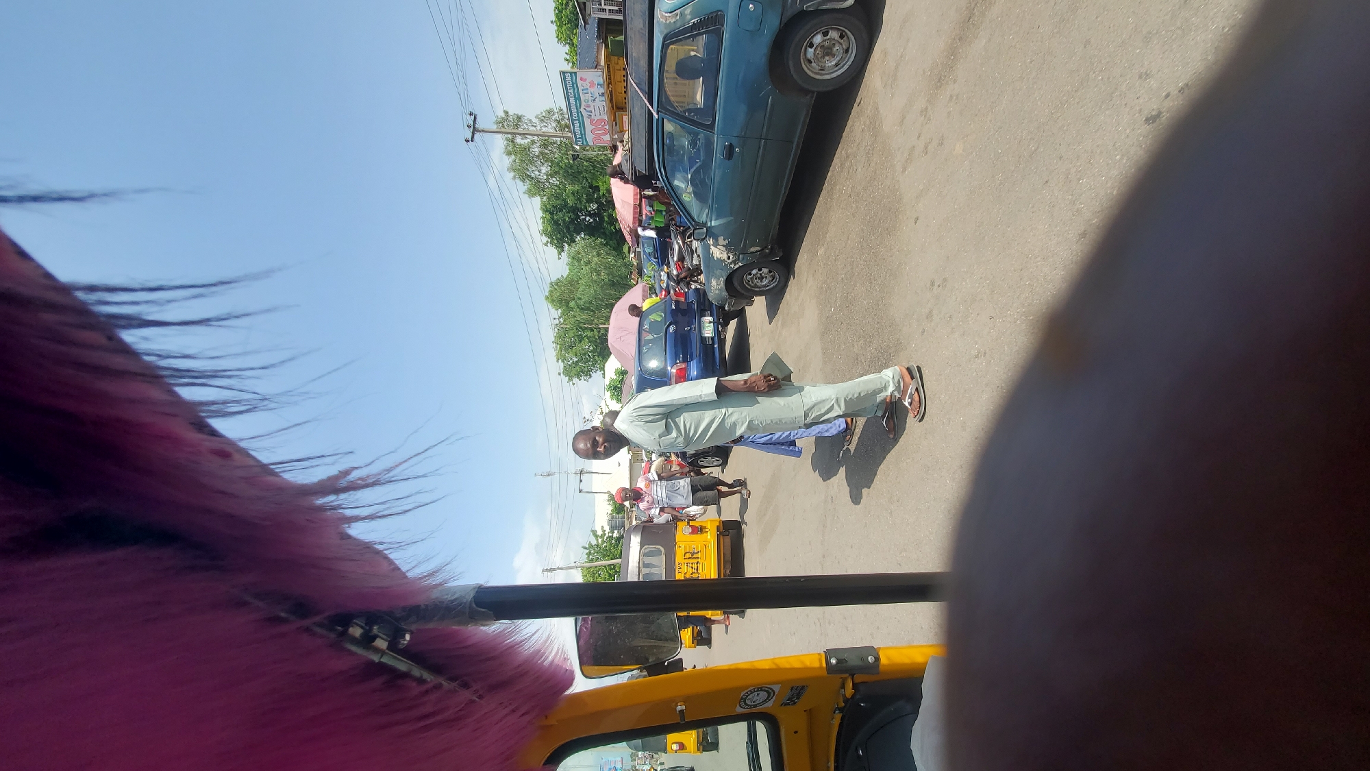 This is a wide-angle, eye-level shot taken on a paved street in Jalingo, Nigeria, during daylight hours. The foreground is dominated by a portion of a yellow motorized tricycle, with a dark, out-of-focus area in the bottom half of the frame, possibly a hand or finger. In the midground, a man in a light green traditional outfit and sandals stands facing the camera, holding a paper in his right hand. Behind him, several vehicles are visible, including a blue hatchback and a turquoise hatchback. A yellow motorized tricycle with an open passenger area is parked to the left. Other individuals are seen in the background, including a man in a red cap and striped shirt, and a person with an umbrella. The environment appears to be a busy street with signs and utility poles in the distance. The sky is clear and blue.