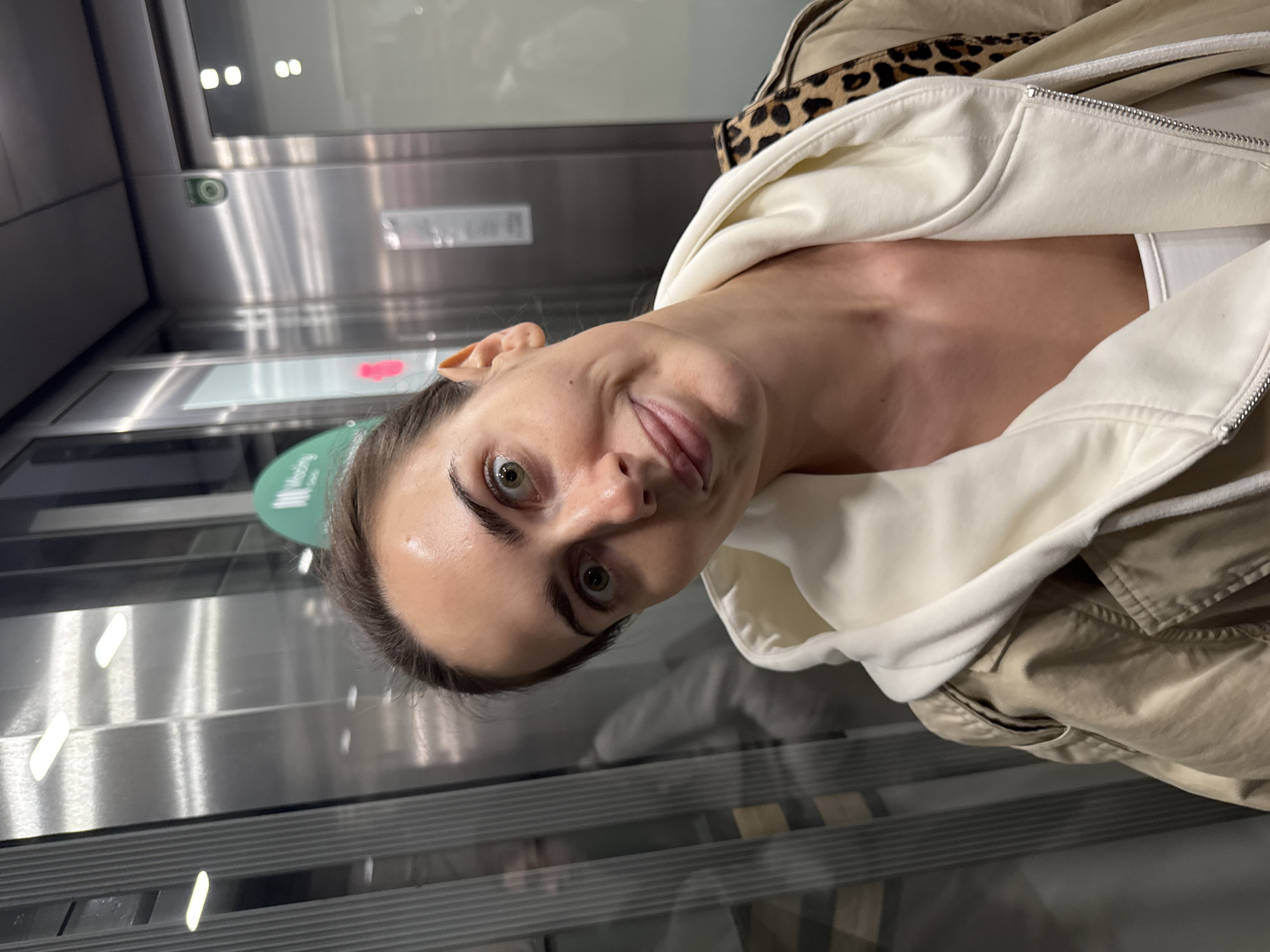 A close-up, selfie-style portrait of a young adult woman taken inside a modern elevator. The image is rotated 90 degrees counter-clockwise, showing the woman's head tilted. She has dark hair, striking green eyes, and a fair complexion, captured with a playful, slightly mischievous expression featuring smirking lips and a hint of her tongue.
She is dressed in a cream-colored hooded sweatshirt or jacket, unzipped, revealing a white top underneath. Over this, she wears a beige or khaki-colored outer jacket, with a leopard print pattern visible on what appears to be the collar or inner lining of the outer garment, near her shoulder.
The setting is an elevator interior, characterized by sleek, reflective stainless steel panels with horizontal detailing. Reflections of overhead lights are visible on the polished surfaces. On the left side of the frame, a green circular indicator with partially visible white text, possibly "Modding" or a similar word, can be seen on the elevator wall. The lighting appears to be artificial, typical of an indoor environment. The photograph captures a casual, lighthearted moment in Warsaw, Poland.