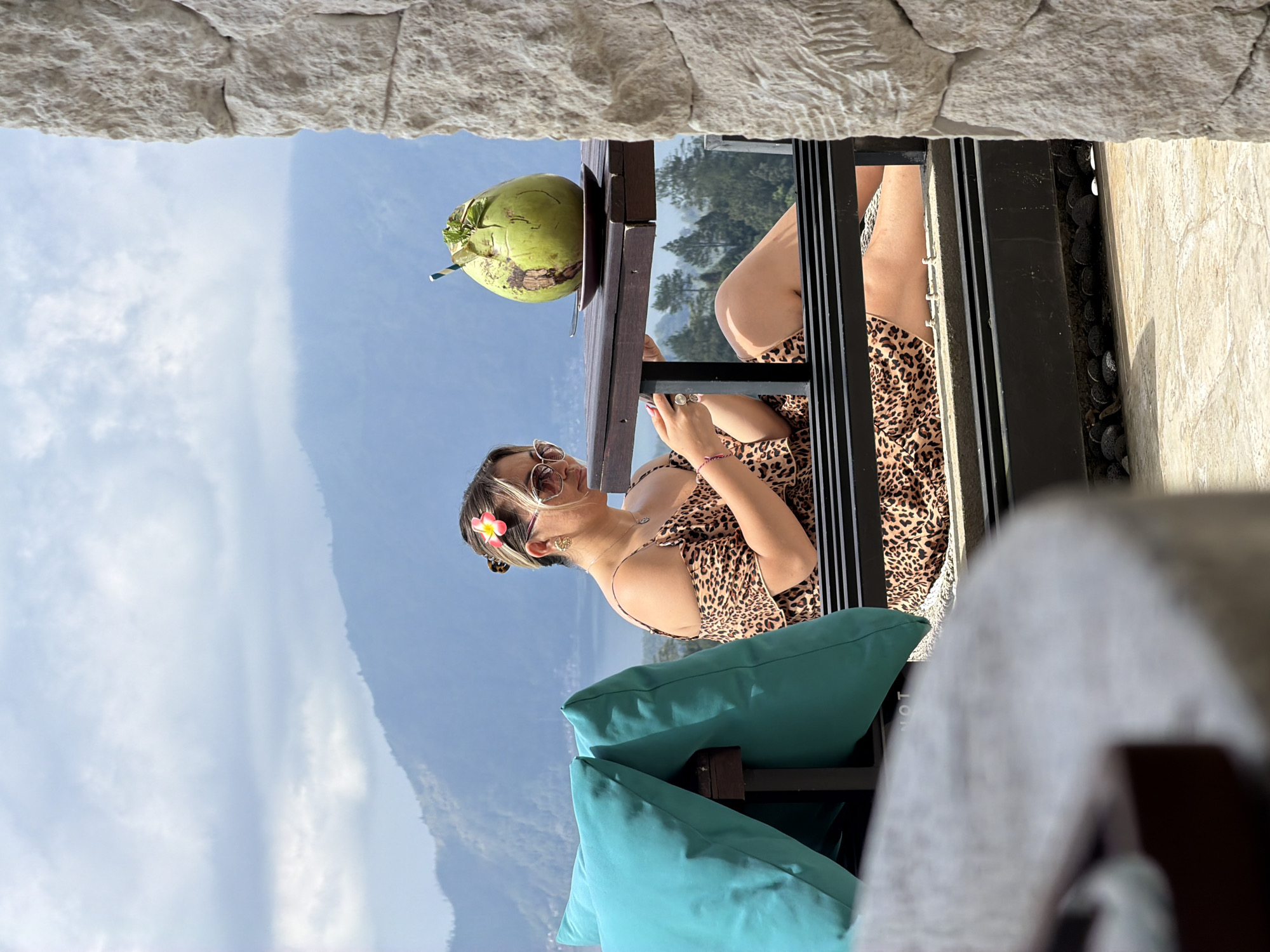 In Kintamani, Indonesia, a woman is seated at a table, looking at her phone. She is wearing a leopard-print dress and sunglasses, with a flower tucked into her hair. A large green coconut with a straw is on the table in front of her. The background shows a scenic view of a lake and mountains under a cloudy sky. The foreground features a stone wall and a chair with teal cushions.