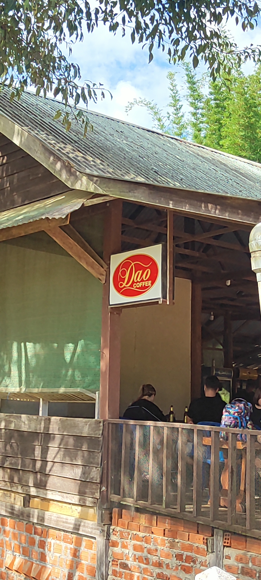 The image shows the exterior of a rustic cafe with a sign that reads "Dao COFFEE". The cafe appears to be open-air, with a wooden structure and a corrugated metal roof. Green netting is visible on the left side, likely providing shade. Several people are seated on a wooden deck in front of the cafe, enjoying drinks. One person is wearing a backpack with a colorful, abstract pattern. The scene suggests a casual, relaxed atmosphere during the daytime, possibly on a warm and sunny day given the bright sky visible through the trees. The architecture and natural surroundings hint at a location in a tropical or subtropical region, consistent with Xayden, Laos. The overall impression is one of a pleasant outdoor dining or coffee-drinking experience.