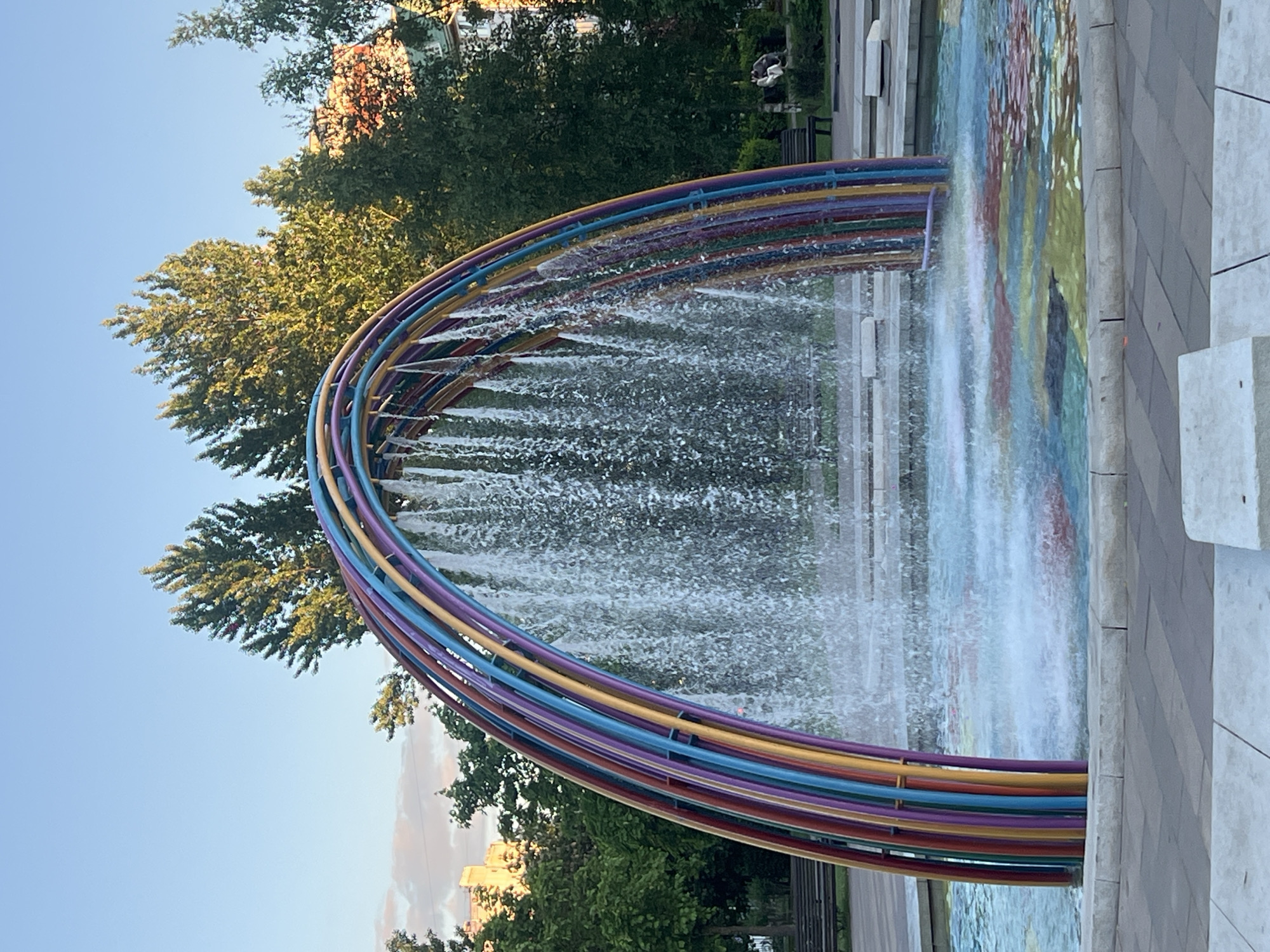 The image depicts a colorful, arch-shaped fountain in Moscow, Russia. The fountain consists of multiple parallel metal pipes, each painted in a different color of the rainbow. Water is cascading from the pipes, creating a curtain effect. The fountain is surrounded by trees and greenery, with a building visible in the background. The sky is clear and blue. A paved area is adjacent to the fountain.