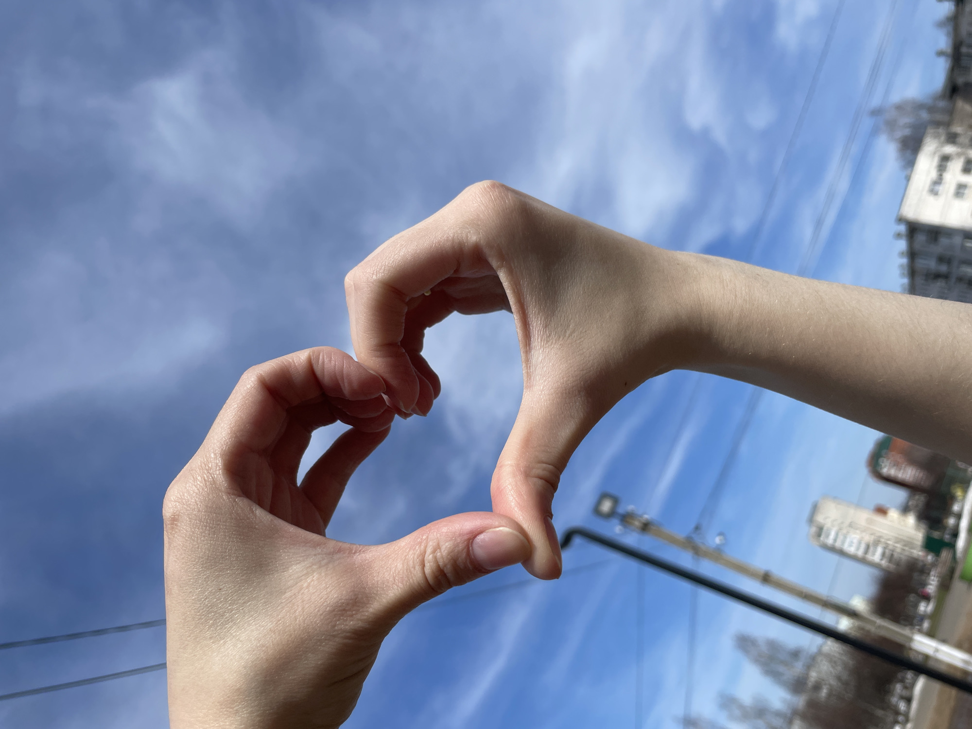 The image features a person making a heart shape with their hands against a backdrop of a cloudy blue sky. The background reveals buildings and power lines, indicating an urban setting. Based on the buildings and other clues, it is possible that this image was taken in the city of Chaikovsky, Russia. However, confirmation of the location would require additional data. The primary subjects are the person's hands and the sky. The action is the hand forming the heart shape. The environment is an urban landscape under a cloudy sky.