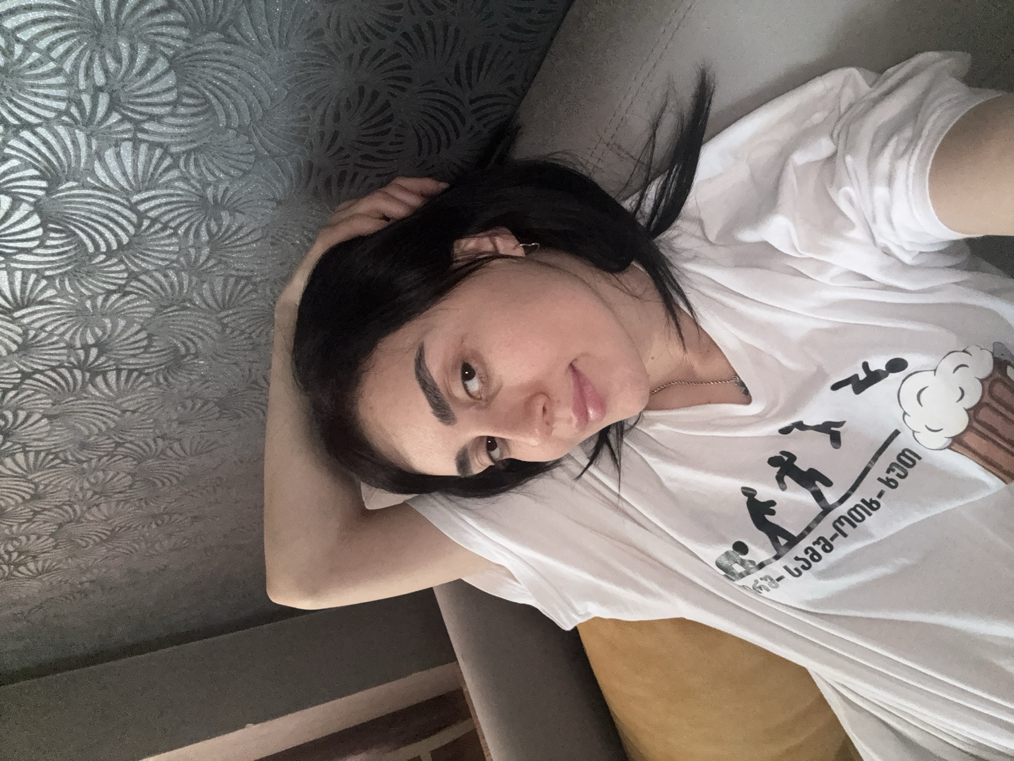 A woman with dark hair is reclining on a grey sofa with a yellow pillow. She is wearing a white T-shirt with a black graphic that includes stick figures and Georgian text. The wall behind her has a grey patterned wallpaper. The location appears to be indoors, possibly a residence. The city of Tbilisi, Georgia is suggested by the Georgian text on the woman's T-shirt.