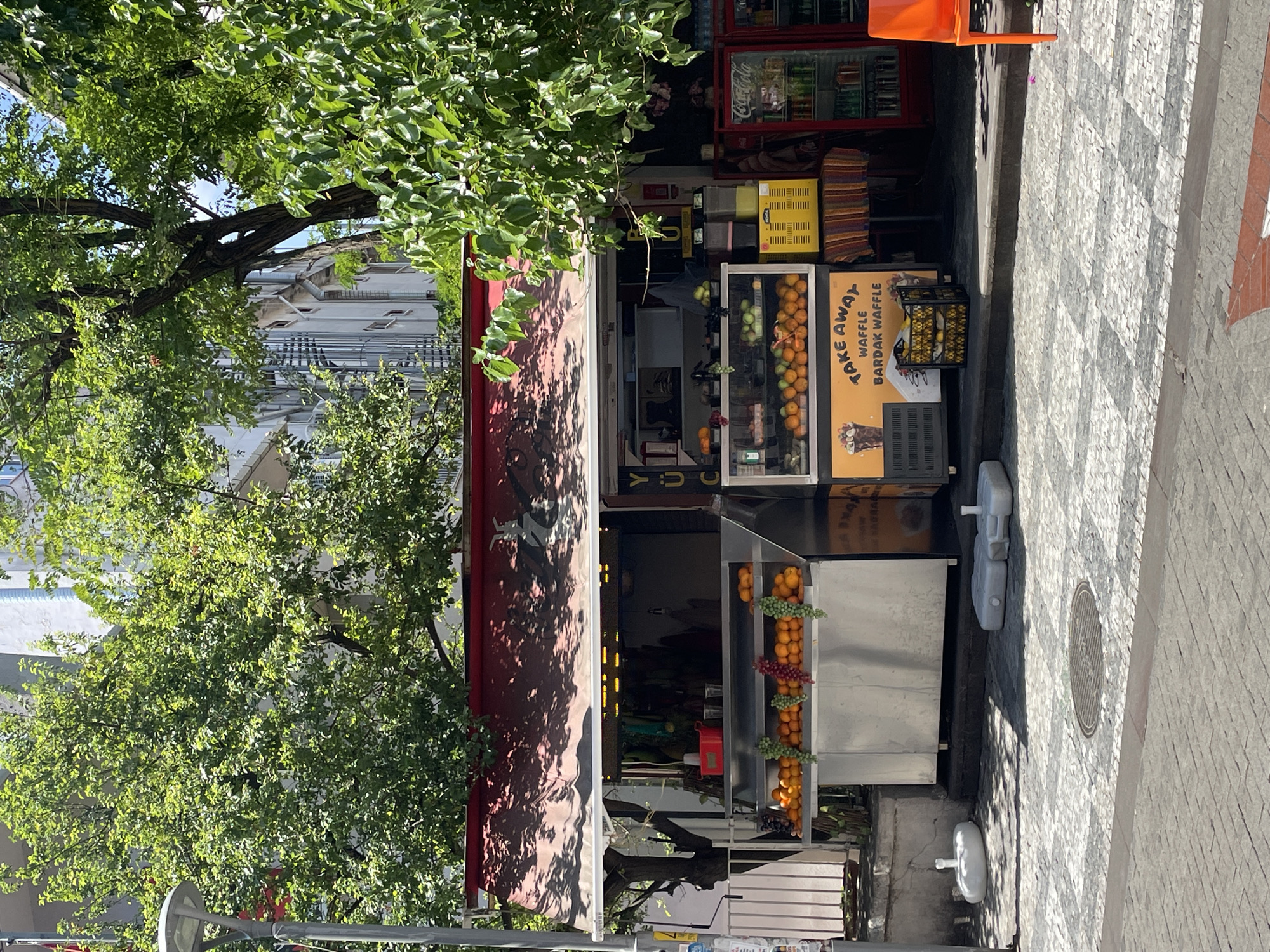 This image depicts a street-level fruit and juice stand located in Kadıköy, Turkey. The stand features a display of fresh fruits, including oranges and grapes, arranged in neat rows. Behind the fruit display, there is a refrigerated glass case showcasing more produce. Above this, a red awning shades the stand, with some signage visible. To the right of the fruit stand, a Coca-Cola branded refrigerator is stocked with beverages. The stand is situated on a cobblestone sidewalk. In the foreground, the cobblestone pattern transitions from a checkerboard design to a more linear arrangement. Two white, water-filled bases for outdoor umbrellas are present on the sidewalk to the left, and one grey, water-filled base is on the ground in front of the stand. In the background, lush green trees overhang the scene, and modern buildings are visible behind the foliage. An orange plastic chair is positioned to the far right of the image.