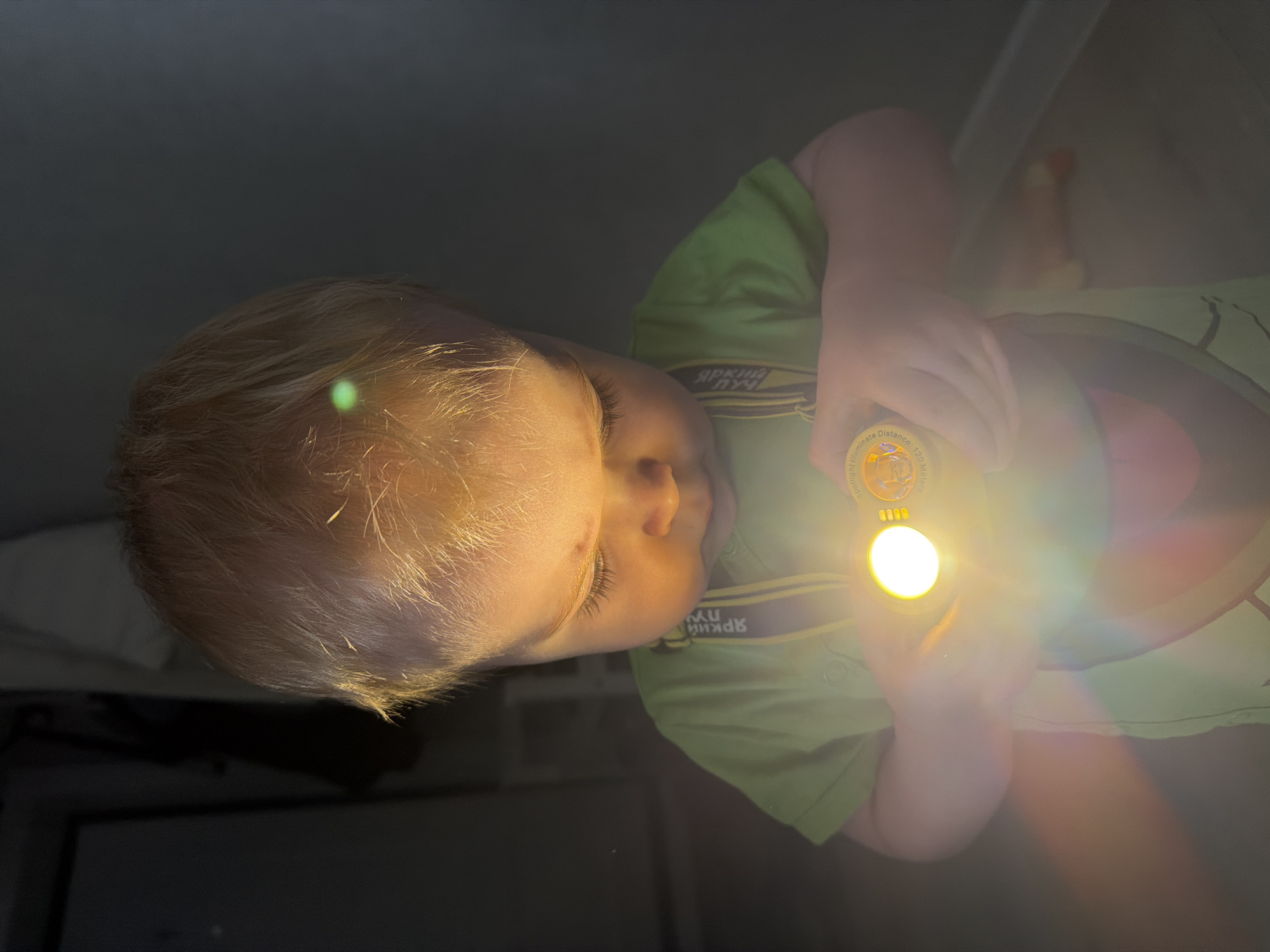 A young, light-haired child wearing a green shirt is holding a lit flashlight in a darkened indoor space. The child's face is illuminated by the flashlight, while the surrounding environment appears dark. The shirt has lettering "Яркий луч" on it. The text on the flashlight mentions "Spotlight Illuminate" and "Distance: 120 meters". The photo does not provide enough information to place the scene within Votkinsk, Russia.
