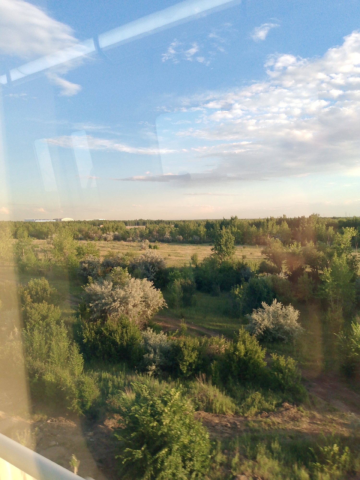 This is a landscape photograph taken from inside a vehicle, likely a train or car, looking out through a window. The foreground features a reflective surface, which is the window itself, obscuring part of the view. Beyond the window, a vast, green landscape unfolds under a bright, partly cloudy sky.
The scene is dominated by lush vegetation, including a variety of trees and shrubs. In the mid-ground, there are open fields dotted with patches of trees and bushes. Further in the distance, a dense line of trees forms the horizon, and beyond that, a large, industrial-looking building complex with white roofs is visible, possibly a factory or a large distribution center. The sky is a clear blue with scattered white clouds, suggesting daytime with fair weather. The light indicates it might be late afternoon or early evening, with a warm, soft glow illuminating the landscape. The overall impression is one of natural beauty interspersed with signs of human development. There is no visible text in the image.