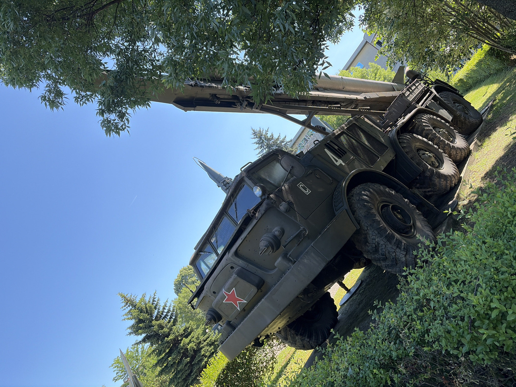 This is an outdoor, eye-level, slightly tilted, waist-up shot of a military vehicle with a missile mounted on top. The vehicle is dark green and has large, rugged tires. A red star symbol is visible on the side of the vehicle's cabin. The vehicle is situated on a grassy incline, surrounded by lush green bushes and trees. The sky is clear and blue with no clouds. In the background, a modern building with windows is partially visible through the trees. This scene is located at the National Museum of Military History in Sofia, Bulgaria, where the vehicle is displayed as an exhibit. There are no people or animals present in the image.