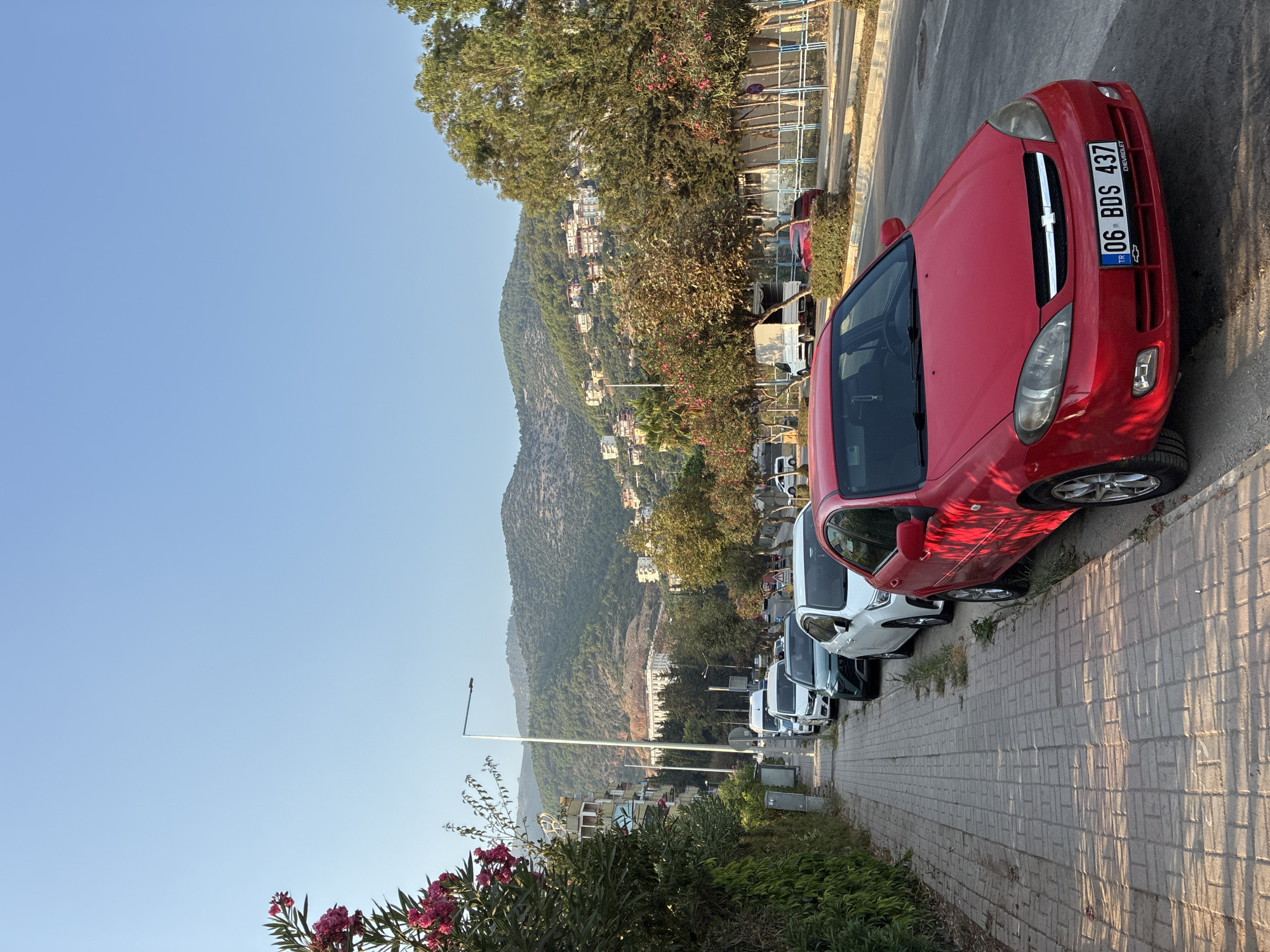 A red Chevrolet sedan is parked on the side of a street in Alanya, Turkey. The car is facing towards the viewer, with its front bumper prominently displayed. Behind the Chevrolet, several other cars are parked in a line along the sidewalk, including a white sedan and a dark green sedan. The sidewalk is made of brick paving stones and is lined with green bushes and trees. In the background, a hillside is visible, dotted with houses and buildings, and a clear blue sky stretches above. The image captures a typical urban street scene with residential architecture and natural elements.