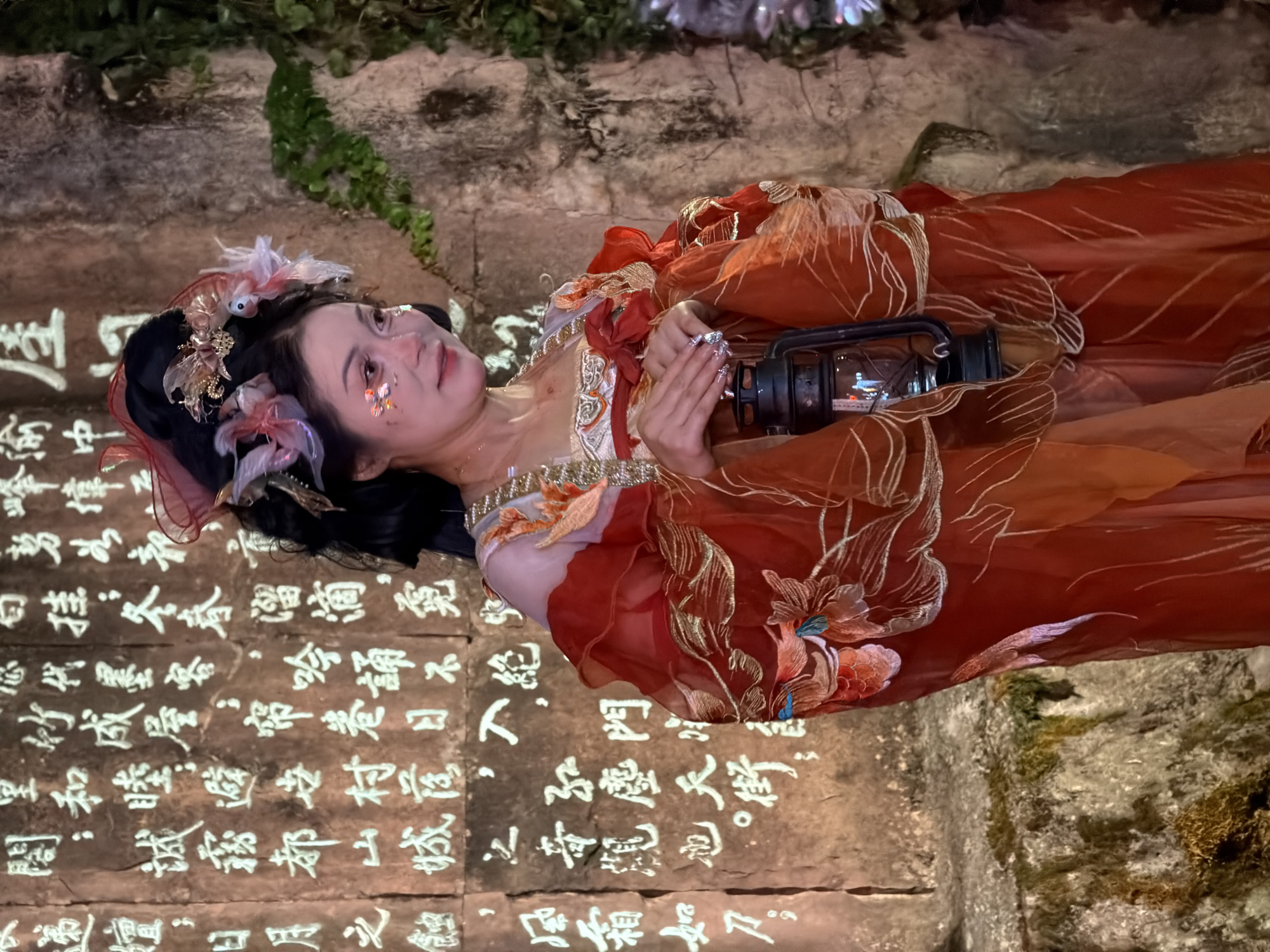 A woman in a red and gold ornate traditional Chinese dress, adorned with floral and butterfly-like ornaments in her hair, holds an old-fashioned oil lantern in her hands. She is positioned in front of a stone wall inscribed with Chinese characters, suggesting a historical or cultural setting. The woman has glitter applied to her face, specifically around her eyes and on her cheekbones. The background shows a rough, moss-covered stone surface and some green foliage. The lighting is dim, focusing on the woman and the illuminated lantern. The scene evokes a sense of mystery and tradition, potentially from a performance or historical reenactment. The Yuzhong District in China is known for its historical sites and cultural heritage, which could be the backdrop for this image.