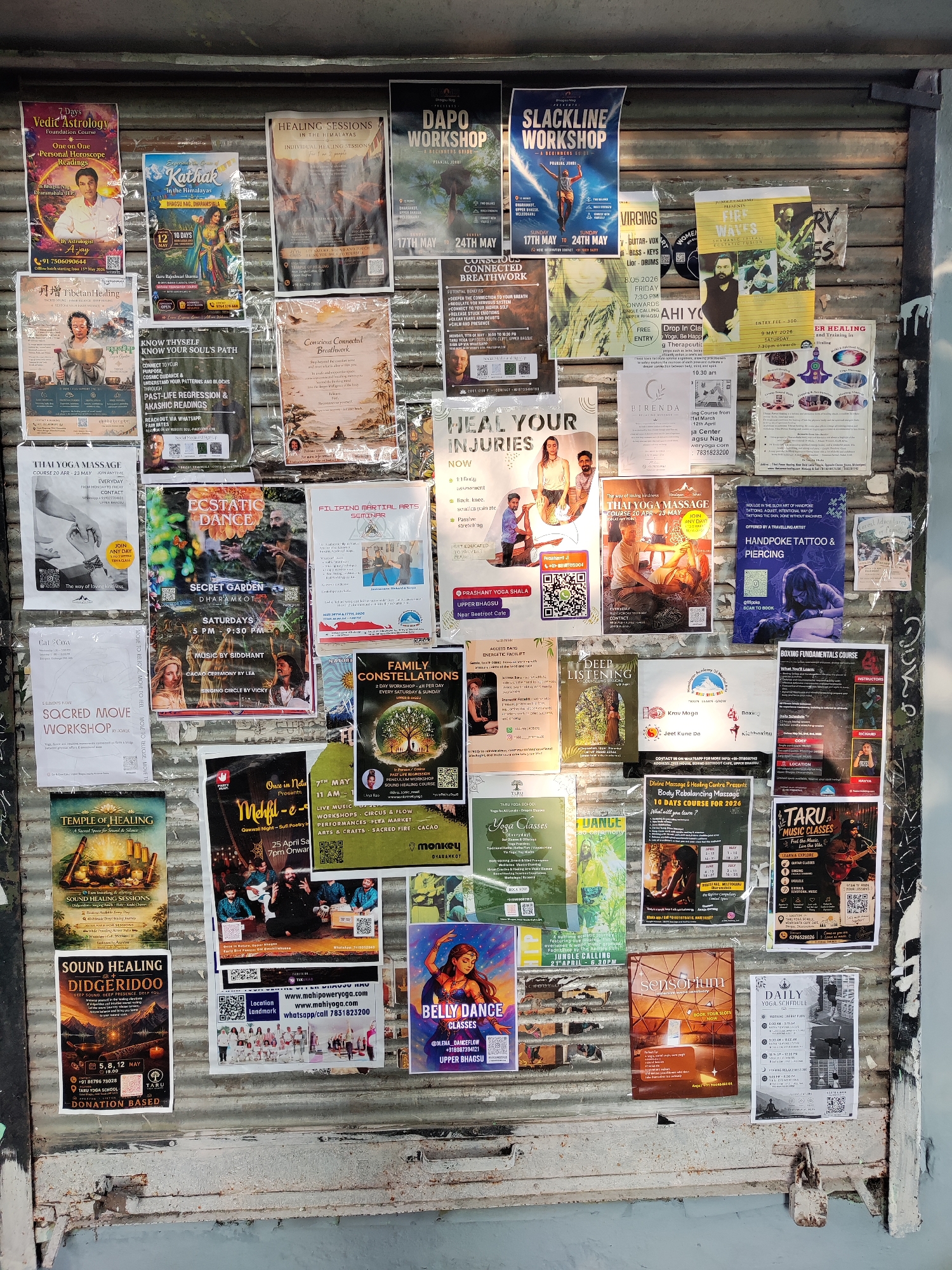 The image depicts a bulletin board on a weathered metal shutter in what is likely a tourist area in India. The board is densely covered with flyers and posters promoting various workshops, healing sessions, yoga classes, music events, and other spiritual or wellness activities. The posters advertise Vedic astrology, Kathak dance, sound healing, Thai massage, ecstatic dance, family constellations, and other alternative practices. The environment is urban, with visible wear and tear on the shutter and surrounding area. The padlock at the bottom suggests the shutter is used to secure a shop or establishment.