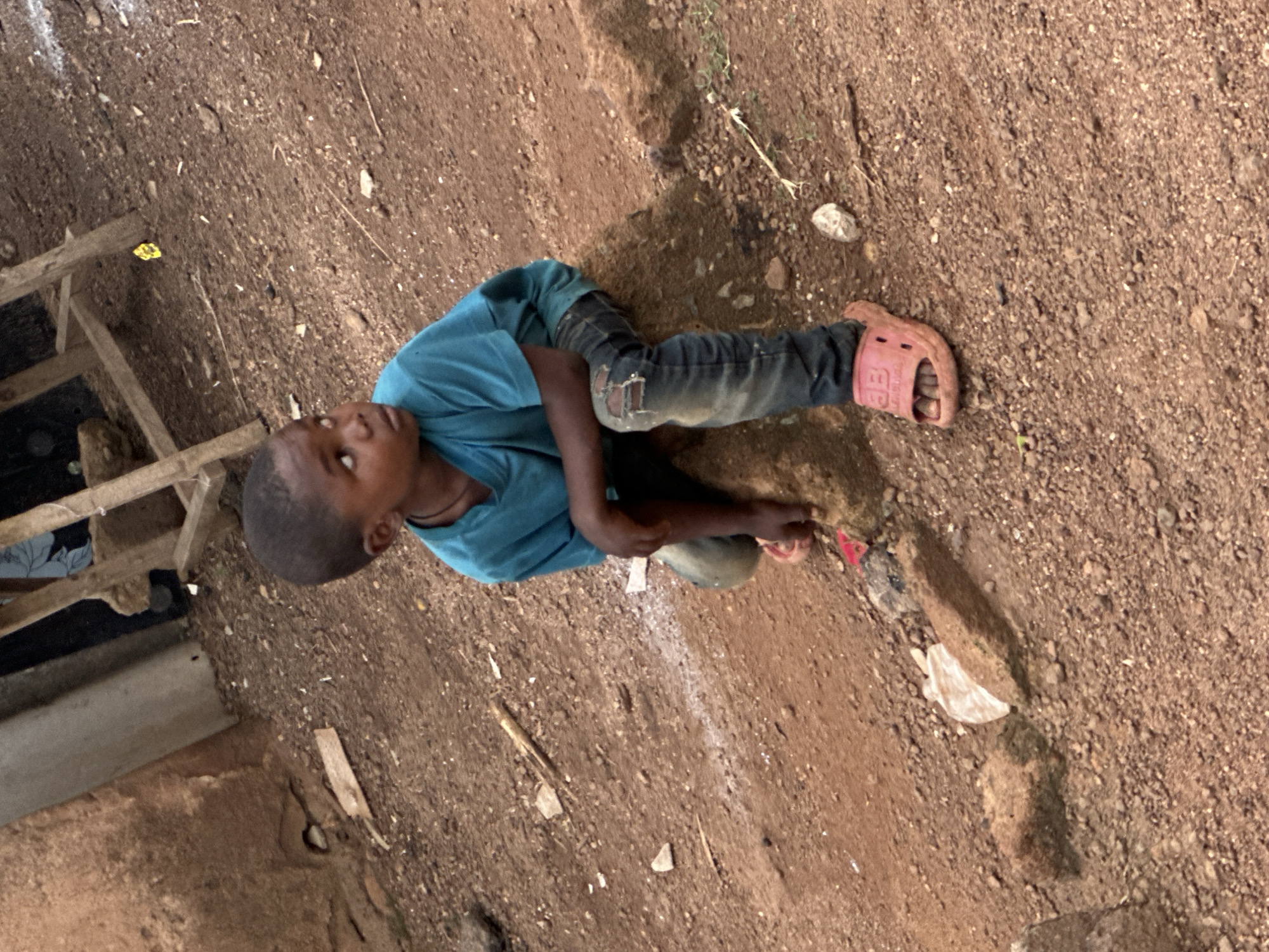 A young boy wearing a teal t-shirt and ripped blue jeans sits on a rock in Gura Top, Nigeria. He is looking to his right, with his hands resting on his knees. The ground around him is made of dirt and small stones, with some scattered debris. In the background, there is a rustic wooden structure and a mud-brick wall. The overall environment appears to be rural or impoverished.