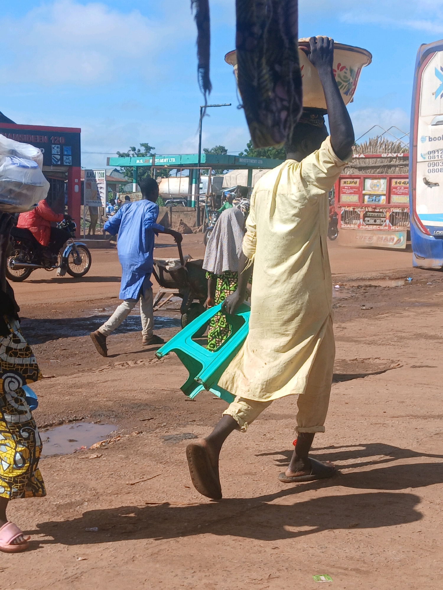 A group of people are walking and pushing a wheelbarrow on a dusty road in Mokwa, Nigeria. The person in the foreground is a man wearing a yellow robe and carrying a yellow bowl on his head. He is walking towards the right side of the frame. A second man, wearing a blue robe and grey pants, is pushing a wheelbarrow to the left. A woman in a colorful patterned skirt and a grey and white patterned headscarf is walking in the background to the right. A motorcycle is parked to the left of the frame. Several trucks are visible in the background. The sky is blue with some white clouds.