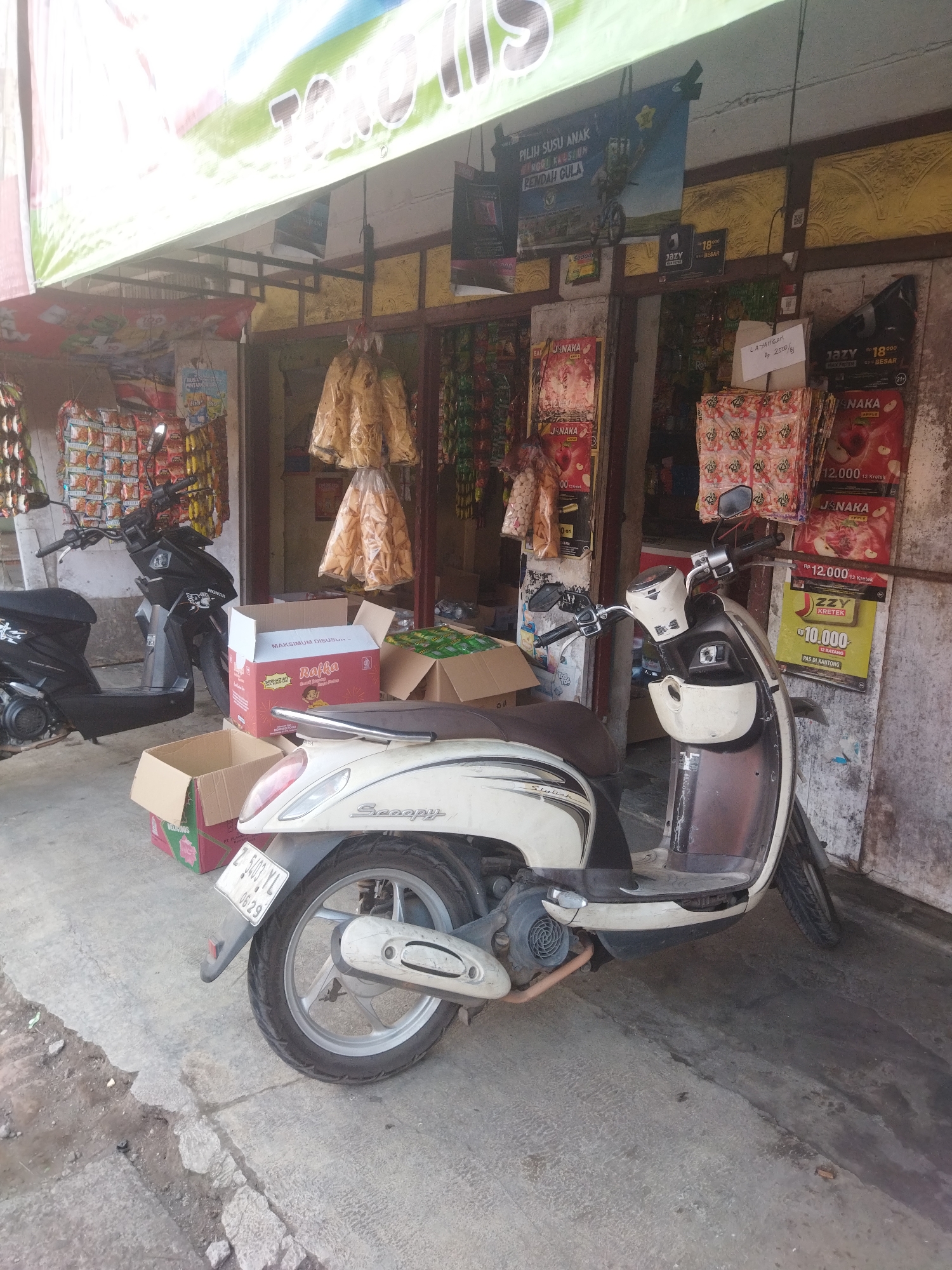 The image shows a street-level view of a small shop in Banjar, Indonesia, likely a warung or convenience store. Two scooters are parked in front of the shop, one black and one white. The white scooter is in the foreground, partially obscuring some of the merchandise displayed outside the shop. The shop itself appears to be selling a variety of packaged goods, including snacks, drinks, and possibly cigarettes, as indicated by the colorful advertisements and hanging displays of products. There are several cardboard boxes in front of the shop, some open, suggesting recent deliveries or stock management. The shop entrance is visible, with a narrow doorway leading inside. The overall scene depicts a typical street-side retail establishment in a bustling Indonesian city.