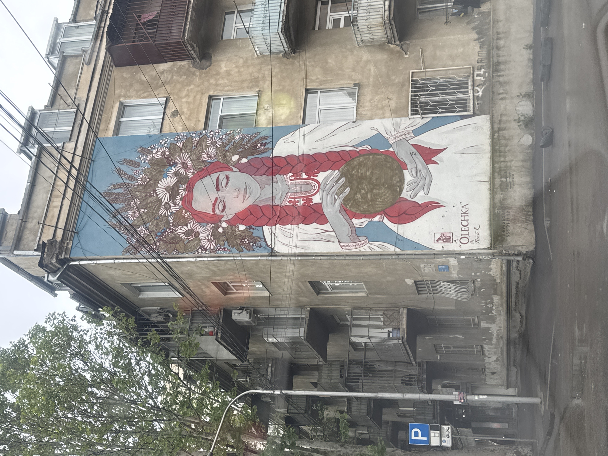 The image shows a street in Tbilisi, Georgia. A large mural depicting a woman with red braids and a floral headdress is painted on the side of a weathered, multi-story building. The woman is holding a brown sphere. The mural includes the text "OLECHKA" and "By Nina L." The building has a number of windows and balconies. Street-level elements include a parking sign, a streetlight, and a tree. The street appears to be wet, possibly from rain.