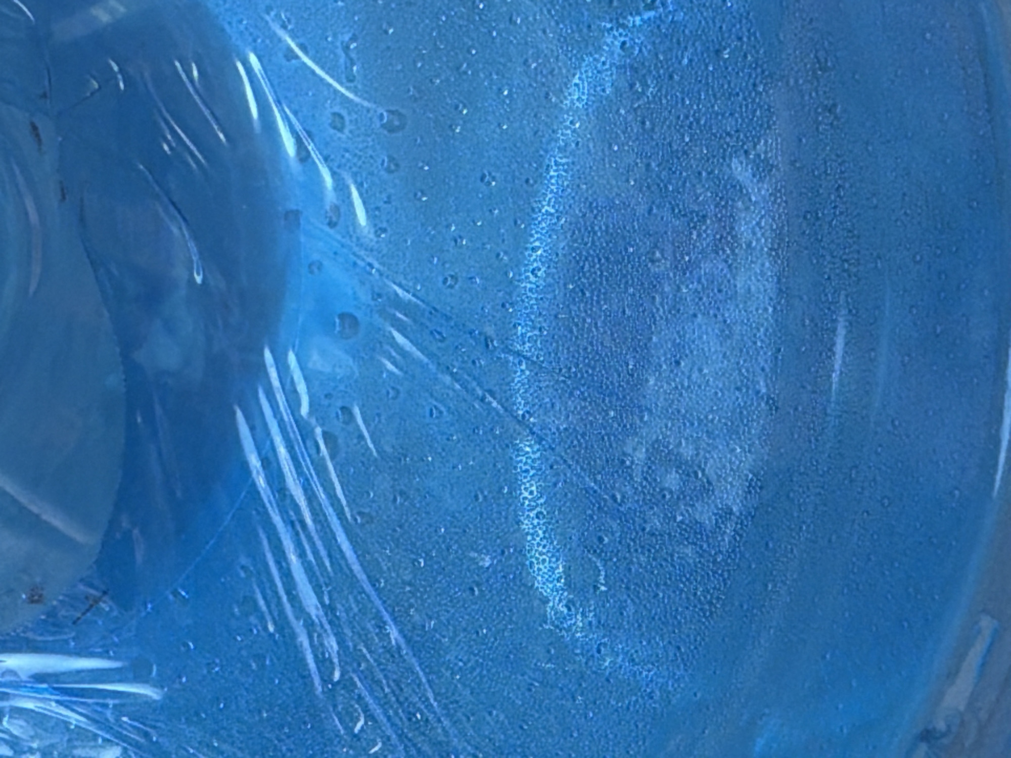 The image shows a close-up, abstract view of a blue plastic water jug with condensation on its surface, covered in plastic wrap. The condensation forms small, numerous droplets, creating a textured appearance on the plastic. The lighting is diffuse, highlighting the blue hue of the jug and the reflective quality of the plastic wrap and droplets. The background is entirely out of focus, consisting of more blue tones and some darker areas, suggesting the jug is in a dimly lit or shadowed environment. There is no indication of the specific location in Nha Trang, Vietnam, as the image focuses solely on the details of the water jug. No people, actions, or interactions are depicted.