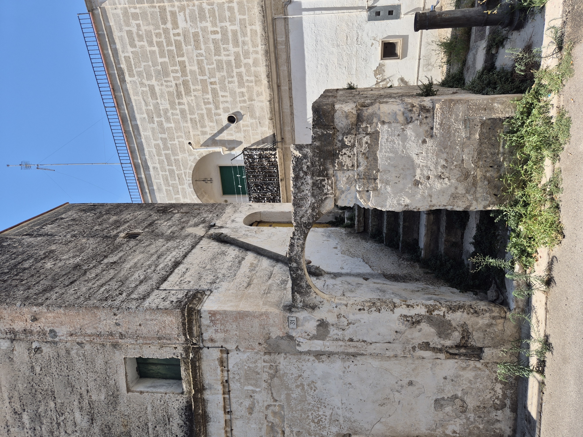 The image captures a scene in Laterza, Italy, focused on the architecture of two adjacent buildings under a clear blue sky. The building on the left is older, constructed with rough, weathered stone and plaster, showing signs of age and decay. It features a small, recessed window with a green shutter and a faded address number "31" above an arched stone passageway. To the right of this passageway, a stone staircase with overgrown vegetation leads upwards.

The building on the right is more contemporary, built with light-colored, uniform brickwork. It has a small balcony with an ornate black metal railing, also featuring a green shuttered window. Above this building, a television antenna is visible against the bright sky, and a black metal railing runs along the rooftop. The ground in the foreground is a paved surface, possibly a street or walkway, with sparse vegetation growing at its edge. A dark, cylindrical object, possibly a water spigot or a post, stands to the right side of the frame, near the base of the brick building. The lighting suggests it is daytime, with harsh sunlight casting distinct shadows.