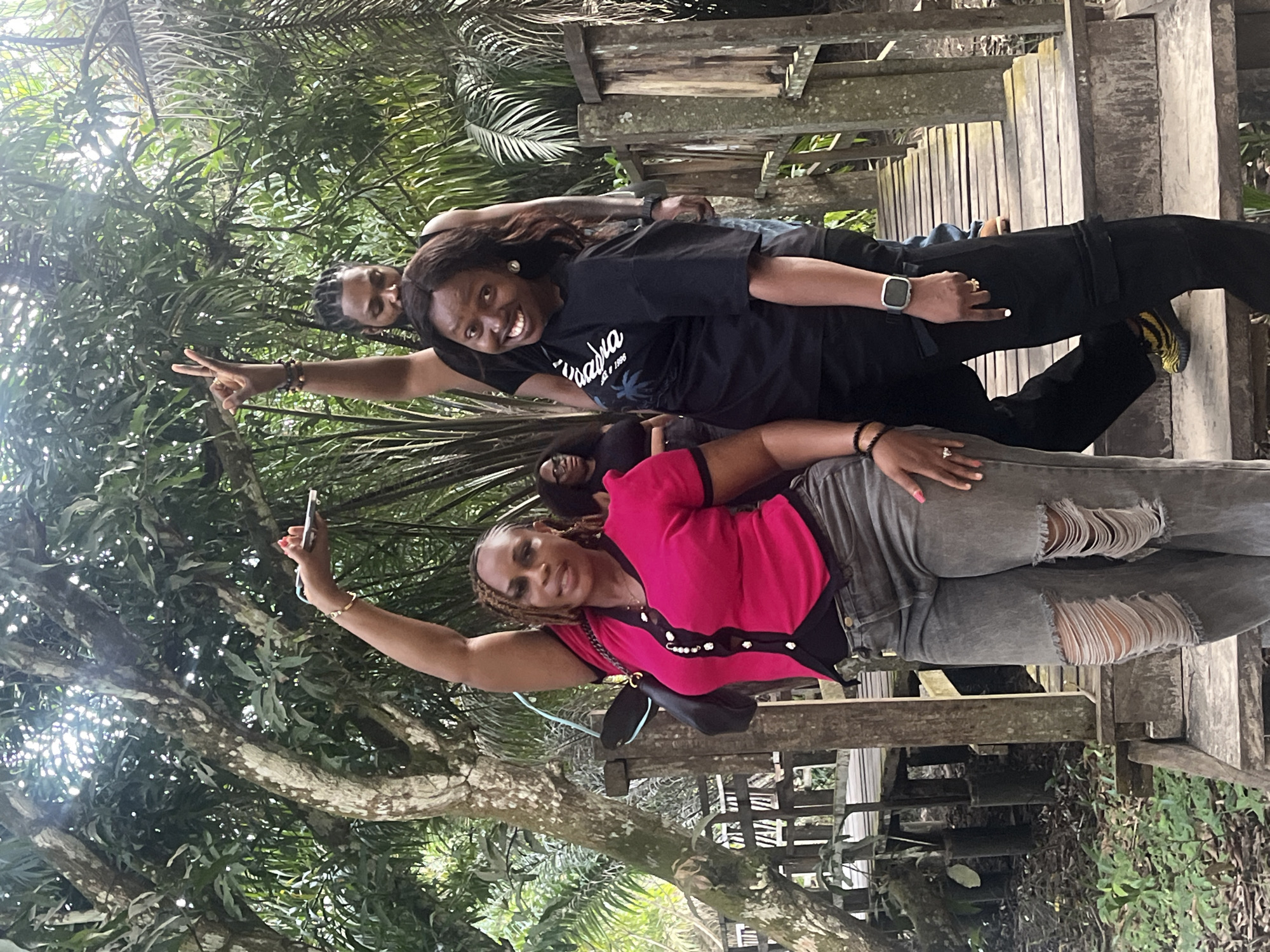 In Lekki, Nigeria, four individuals are gathered on a wooden walkway amidst dense foliage. Two women are in the foreground, one wearing a pink top and ripped grey jeans, striking a pose with her arm raised and hand holding a phone. The other woman, in a black t-shirt and black pants, smiles at the camera, also with an arm raised in a peace sign. Behind them, a man with braided hair, also wearing a black top, has his arm raised in a peace sign. Another woman is partially visible in the background, wearing glasses and a black top. The walkway appears rustic, constructed of weathered wood, and is surrounded by lush green trees and palm fronds, suggesting a natural or park-like setting within Lekki.
