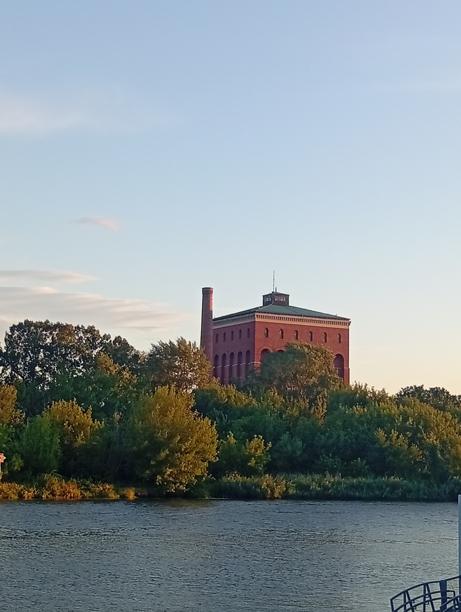 The image depicts a brick building with a tall chimney, situated on a tree-covered island in a river. The building appears to be an old industrial structure, possibly a former factory or power plant. The sun is setting, casting a warm glow on the scene. The river is calm, with gentle ripples on its surface. The sky is clear, with a few wispy clouds. This building is the former insulation factory in Wrocław, Poland, located on Ostrów Tumski.