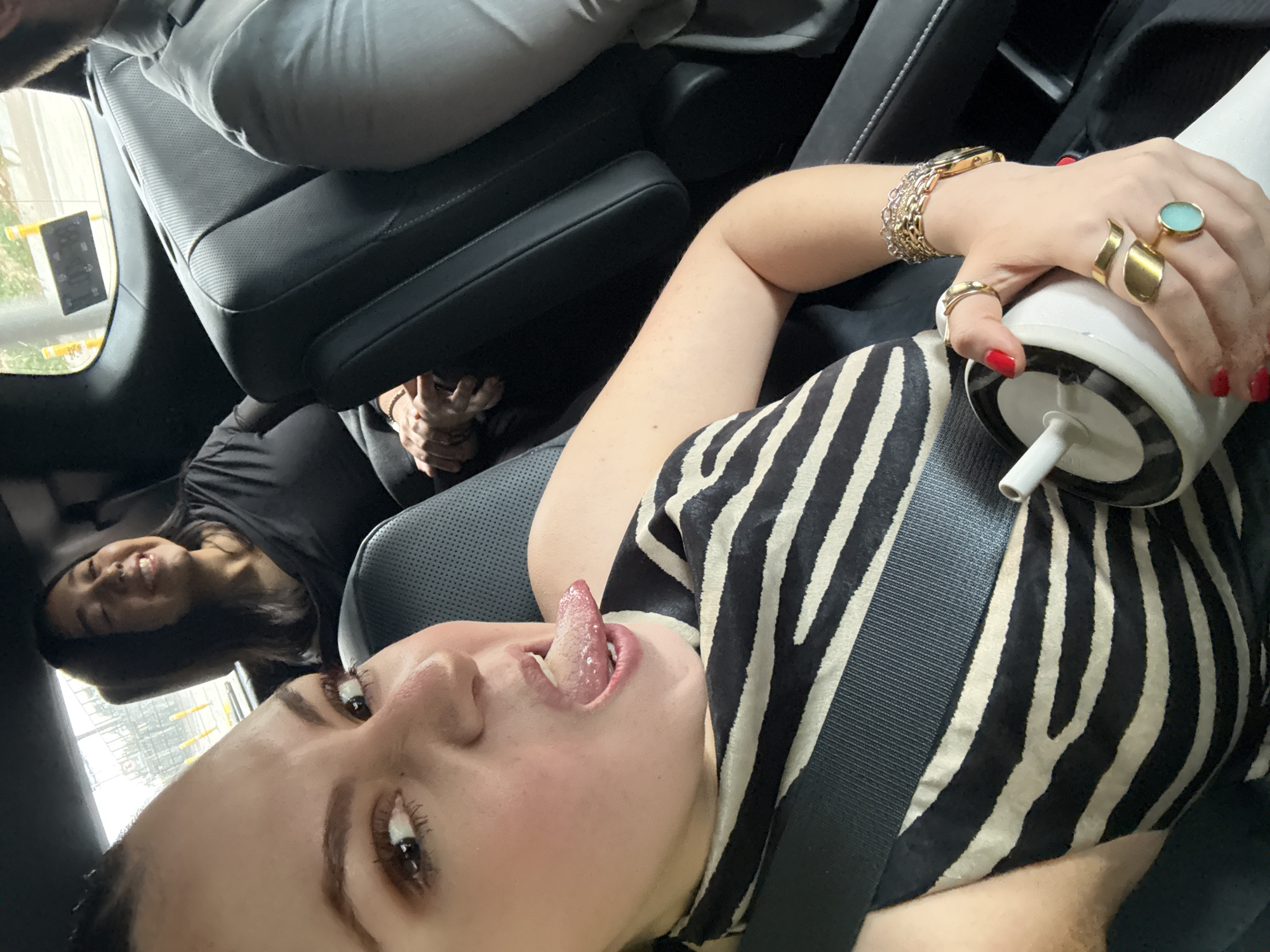 The image appears to have been taken inside a vehicle. There are at least two occupants visible. The primary subject, closest to the camera, is a light-skinned person with dark hair, wearing a zebra-striped shirt and a seatbelt. This person is holding a white cup with a straw and has their tongue sticking out. Their left hand is adorned with multiple gold rings and a gold bracelet.
In the background, another person with dark hair is visible in another seat. There is a person in the front seat. The car's interior is black.
The background visible through the car's window appears to be a street in an urban area, possibly Mexico City, Mexico. Details are limited due to the focus and proximity to the primary subject. There appear to be building exteriors that suggest architecture found in Mexico City, Mexico. However, without clearer external landmarks, a definitive location cannot be confirmed.