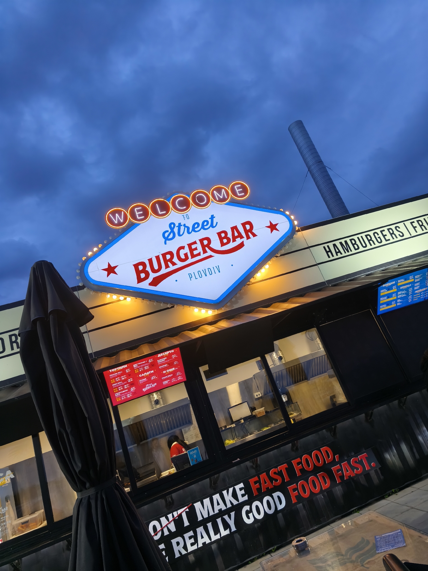 Here's a detailed description of the image:
**Scene:** An outdoor shot of a food establishment called "Street Burger Bar" in Plovdiv, Bulgaria. The image is taken at twilight, as evidenced by the deep blue, cloudy sky.
**Subjects/Objects:**
* **Street Burger Bar:** The main focus. The signage features a "Welcome to" marquee-style display with the name in a star-shaped sign. The word "Plovdiv" is written below.
* **Menu boards:** Digital menu boards are visible in the windows displaying food options and prices.
* **Structure:** The bar appears to be a converted shipping container.
* **Umbrella:** A folded black patio umbrella is located in the left foreground.
* **Tables:** Tables are placed in front of the bar for customers.
* **Chimney-like structure:** A tall, cylindrical structure is located behind the bar.
* **Text on the Bar:** Slogan is visible, written in bold red and white letters. "DON'T MAKE FAST FOOD. REALLY GOOD FOOD FAST."
**Environment:**
* The time of day is dusk.
* Cloudy sky.
* The area appears to be an outdoor dining space.