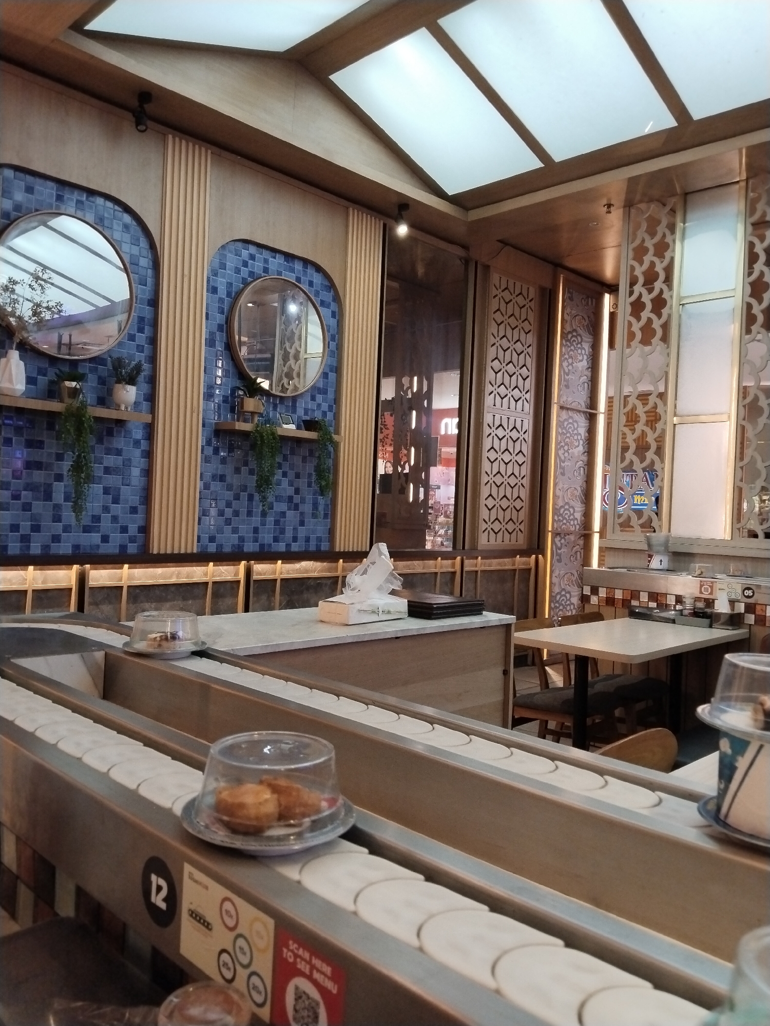 The image shows a conveyor belt sushi restaurant interior. In the foreground, a conveyor belt is visible with plates of food covered by plastic domes. The belt is made of metal and wood. In the background, there are tables and chairs, with decorative panels on the walls. Two round mirrors are mounted on a blue tiled wall. The ceiling has a modern design with multiple light panels. The restaurant appears to be clean and well-lit. The scene does not show any people or specific actions taking place, focusing on the restaurant's ambiance and setup. The location of Kudus, Indonesia is not evident from the image itself, as there are no identifying markers or contextual clues suggesting this specific city.