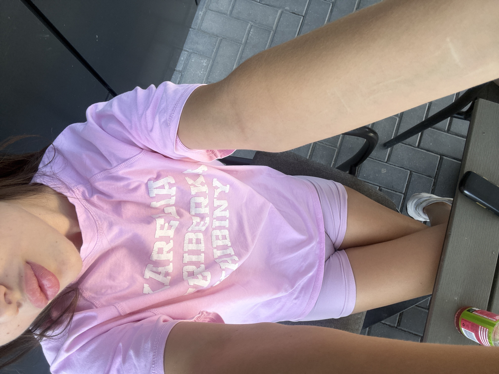The image shows a person wearing a pink t-shirt and shorts. The t-shirt has white lettering on it, which appears to say "KARERA RIBERY" in the top two lines and possibly "HIBINY" on the third line. The person is holding their arm up, and the image is taken from a high angle. In the background, there is a dark surface, possibly a table or umbrella, and a brick-paved area. A can of soda and a phone are visible on a table in the foreground. The scene is outdoors, with natural light. The image does not contain any information that would allow for its location to be determined as Comrat, Moldova.